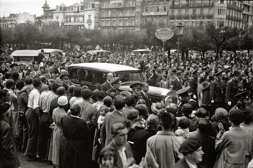 Zdjęcie przedstawia przejeżdżający samochód, który jest otoczony przez tłum ludzi. W tle znajdują się kamienice.