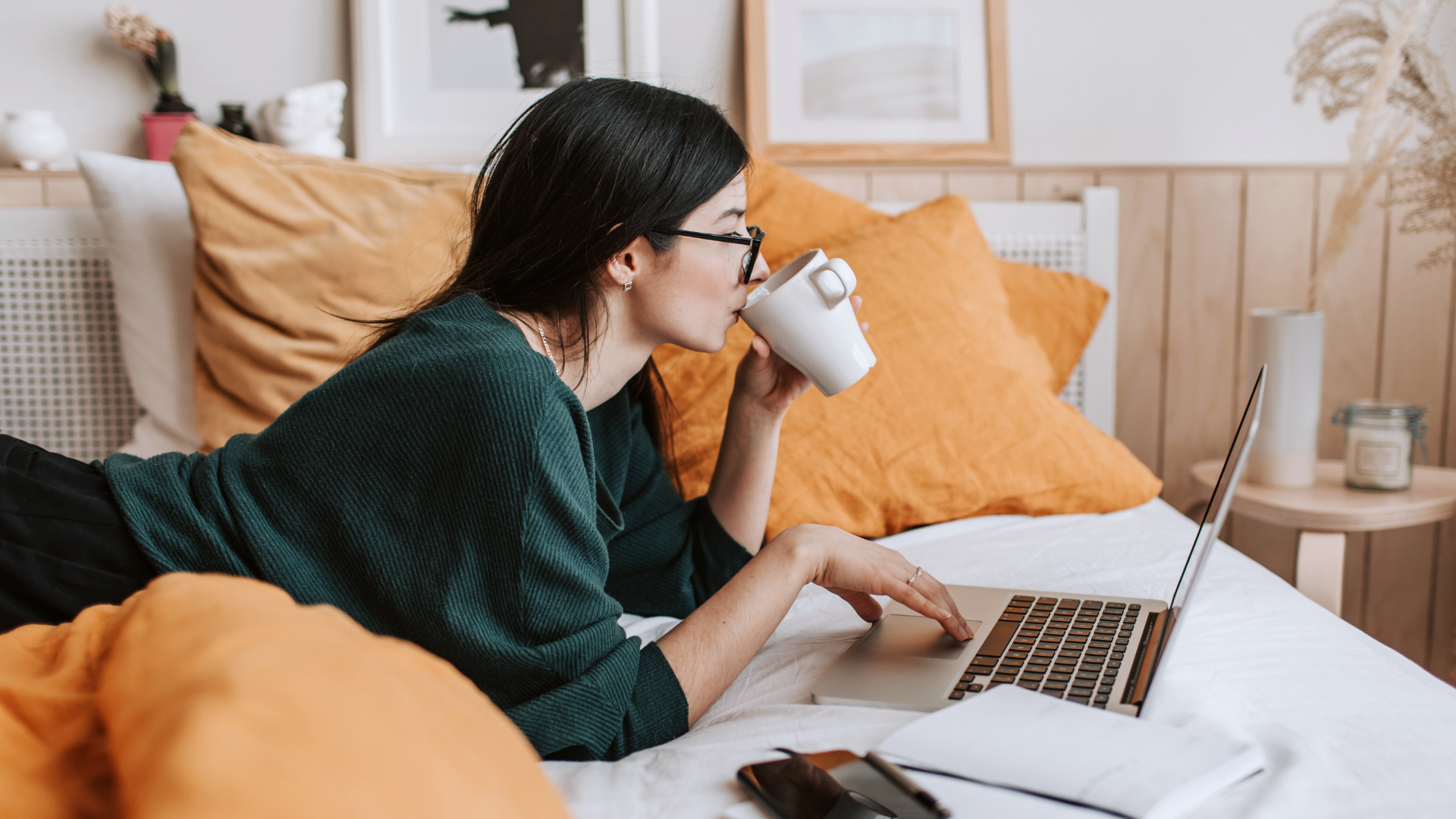Zdjęcie przedstawia dziewczynę w okularach, która leży na brzuchu na łóżku. Przed nią znajduje się laptop. Prawą ręką dotyka touchpada, a lewą zbliża kubek do ust. W tle widoczne są poduszki, stolik przy łóżku, obrazki na ścianie.