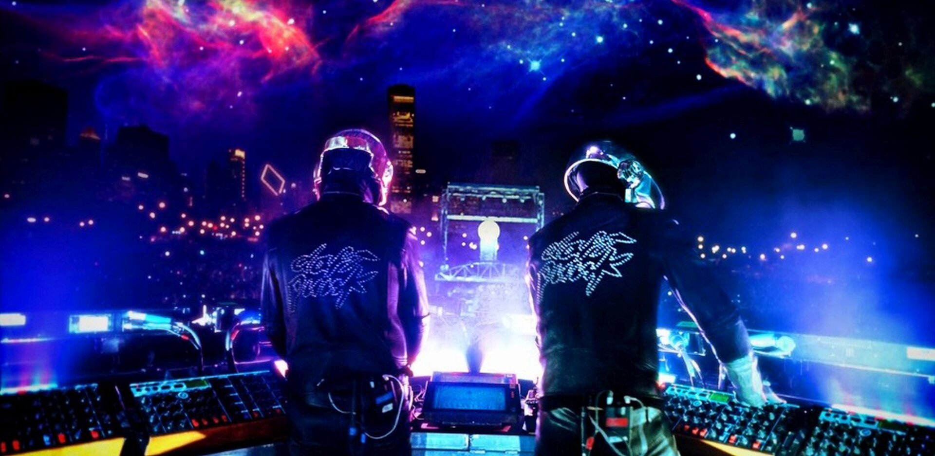 Na zdjęciu dwóch DJ-ów sfotografowanych podczas tworzenia muzyki, od tyłu. Przed mężczyznami konsole. Muzycy ubrani są w czarne stroje z jasnymi elementami, które świecą w świetle ultrafioletowym. W tle pokaz świetlny.