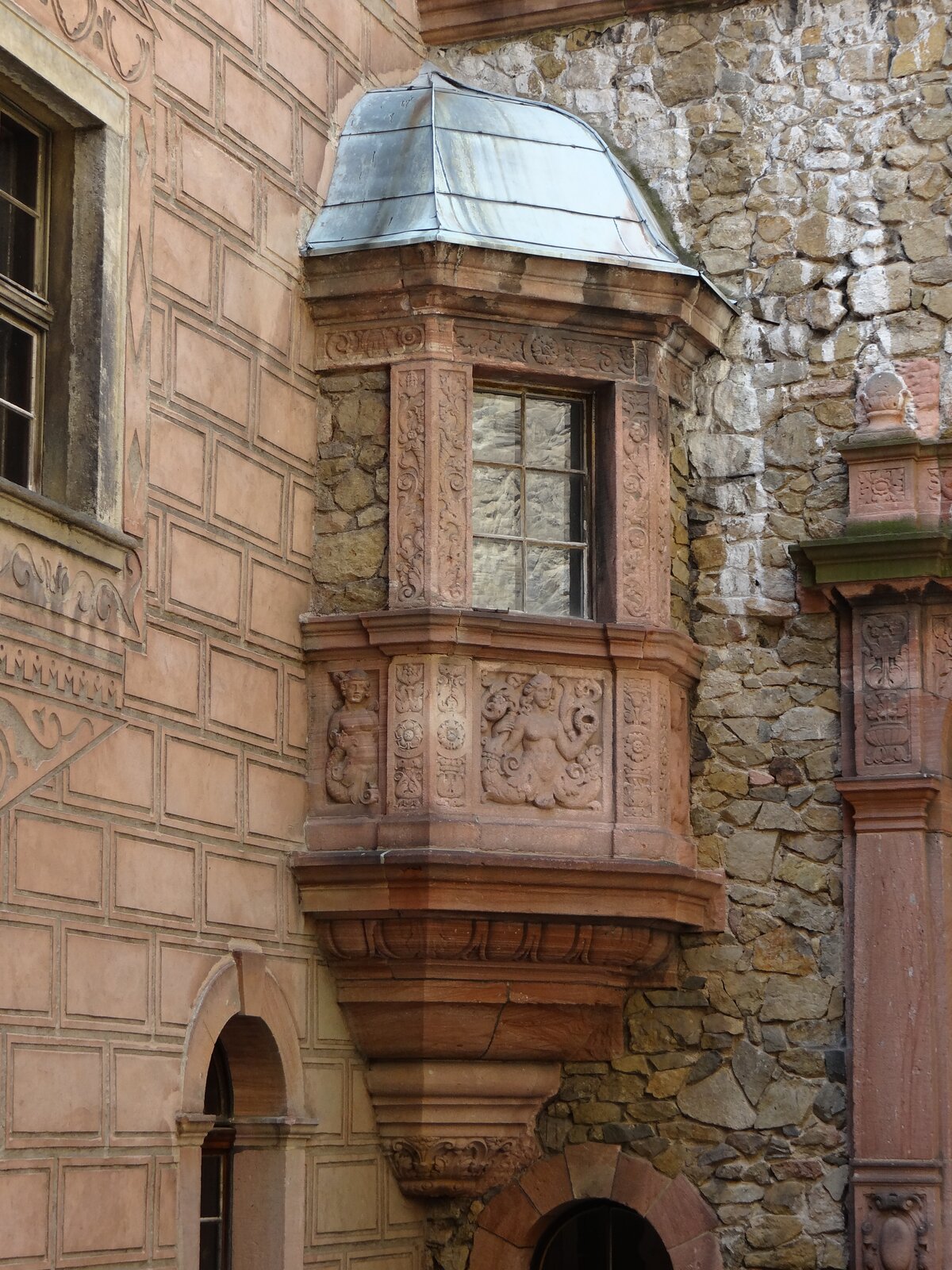 Fotografia przedstawia element architektoniczny - wykusz, który wystaje poza elewację budynku - mury tego elementu są bogato zdobione reliefami, które przedstawiają nagie kobiety. Wykusz posiada jedno okno metalowy dach w srebrnym kolorze. Zamocowany jest pomiędzy dwoma ścianami budynku.