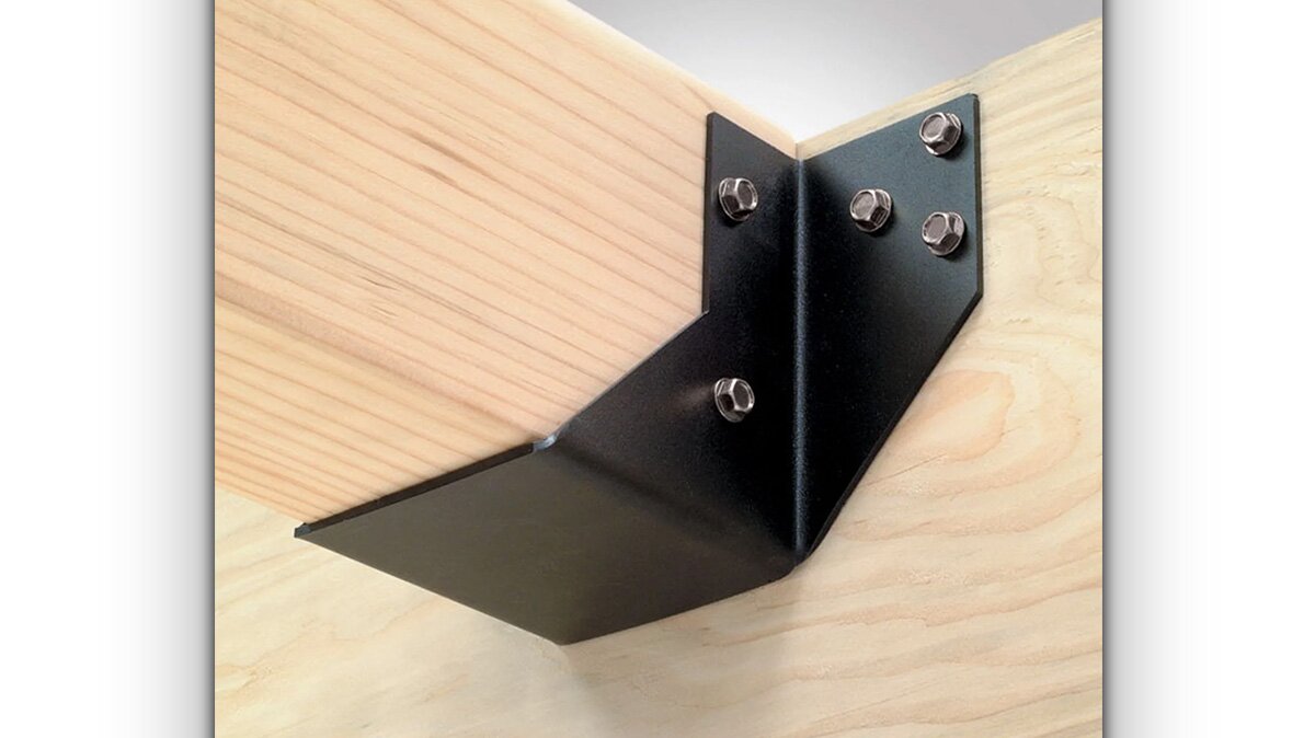 Na zdjęciu widoczny punkt styku dwóch belek, które znajdują się prostopadle do siebie. Belki te są ze sobą złączone czarną blaszką z wypustkami, która tworzy kąt prosty pomiędzy belkami, a także zachodzi na dół jednej z nich, wzmacniając łączenie.