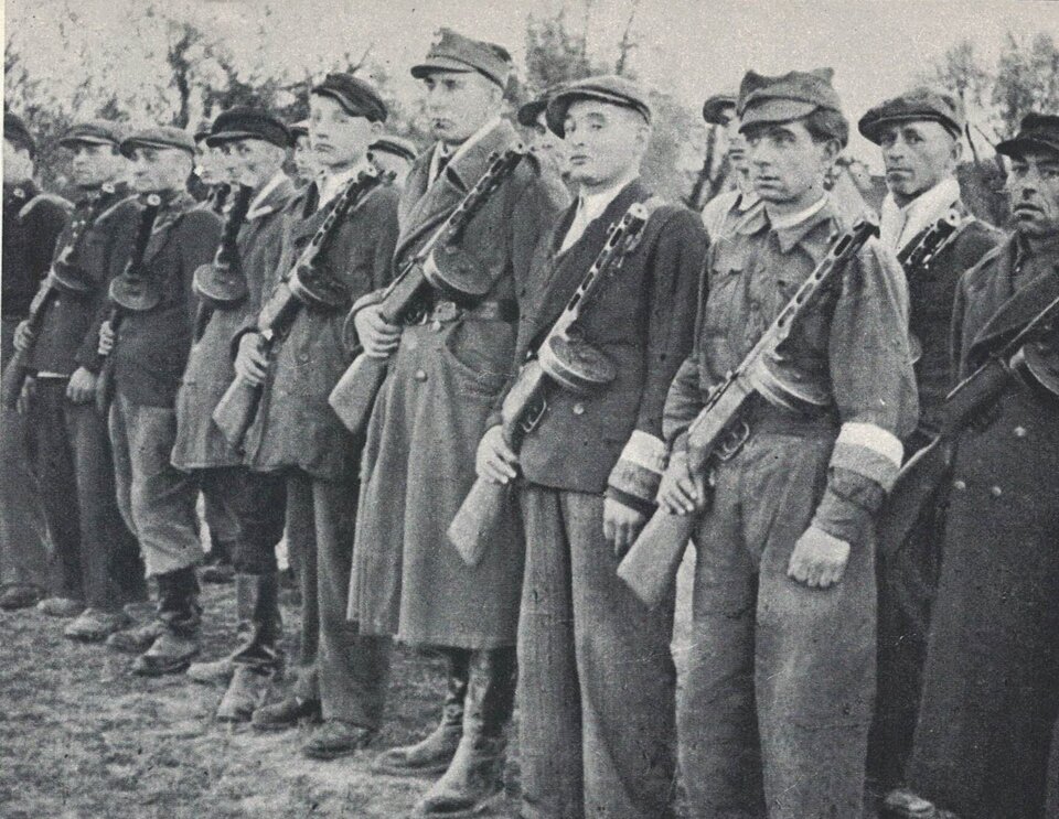 Zdjęcie przedstawia kilkunastu mężczyzn ustawionych w dwóch równych szeregach, którzy pozują do zdjęcia. Są oni w różnym wieku, zarówno młodzi jak i starzy. Niektórzy z nich ubrani są w mundury lub płaszcze wojskowe i czapki rogatywki, inni mają stroje cywilne: marynarki, spodnie, koszule i płaskie czapki z daszkiem. Buty są krótkie lub długie z cholewą.  Mężczyźni są uzbrojeni w karabiny maszynowe, które opierają na lewym ramieniu. Niektórzy z nich na lewym przedramieniu mają opaski z biało‑czerwoną flagą.   