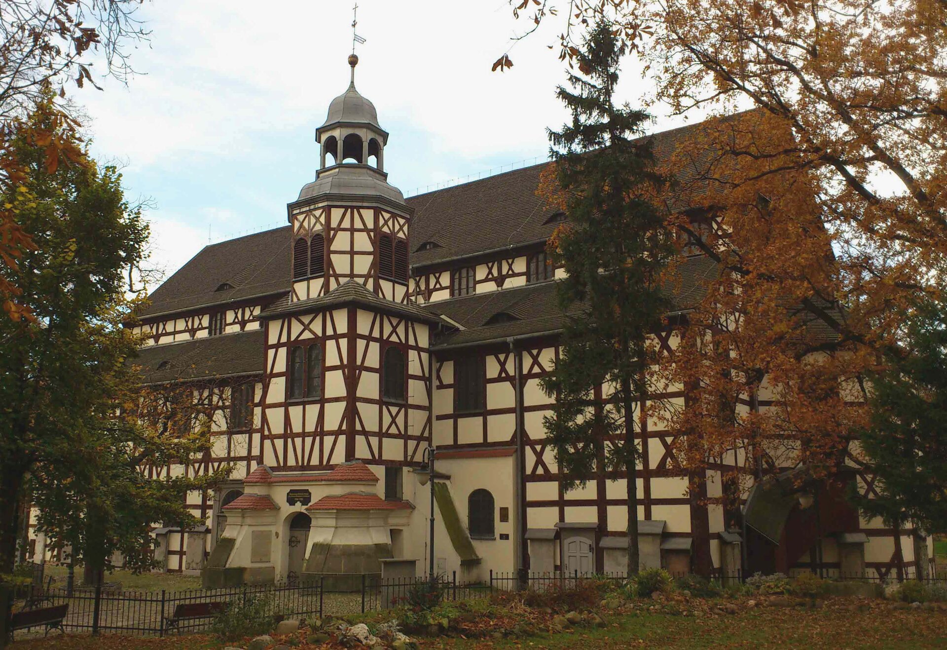 Na zdjęciu protestancki kościół o konstrukcji szachulcowej – ściana szkieletowa drewniana (ciemne belki ułożone w kratkę i skos), wypełnienie stanowi glina otynkowana na kolor piaskowy. Dach skośny brązowy, jedna wieża znajdująca się na froncie, w niej wejście. Dookoła budynku niski płot, teren porośnięty roślinnością. 