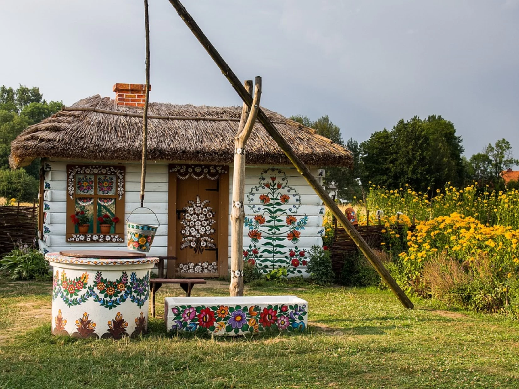 Zdjęcie przedstawia mały, biały domek zdobiony malowidłami, ustawiony na łonie natury. Ściany wykonane są z drewnianych bal, pomalowanych na biało. Z przodu znajduje się drewniane okno oraz drewniane drzwi. Okno jest zdobione malowidłami z kwiatowymi motywami w kolorach białym oraz zielonym, żółtym i różowym. Okno jest dzielone na cztery drewnianymi belkami. Na parapecie stoją dwie doniczki z czerwonymi kwiatami. Drzwi są wysokie do samego dachu. Na nich również są białe kwiatowe motywy. Na ścianie po prawej stronie drzwi namalowano duży obrazek. Dach jest zrobiony ze strzechy. Z niego wystaje murowany komin. Przed domkiem, wśród trawy stoi studnia z drewnianym żurawiem. Studnia oraz wiadro są zdobione kwiatowymi malunkami. Po prawej stronie domu rosną wysokie, żółte kwiaty. W tle rosną drzewa.