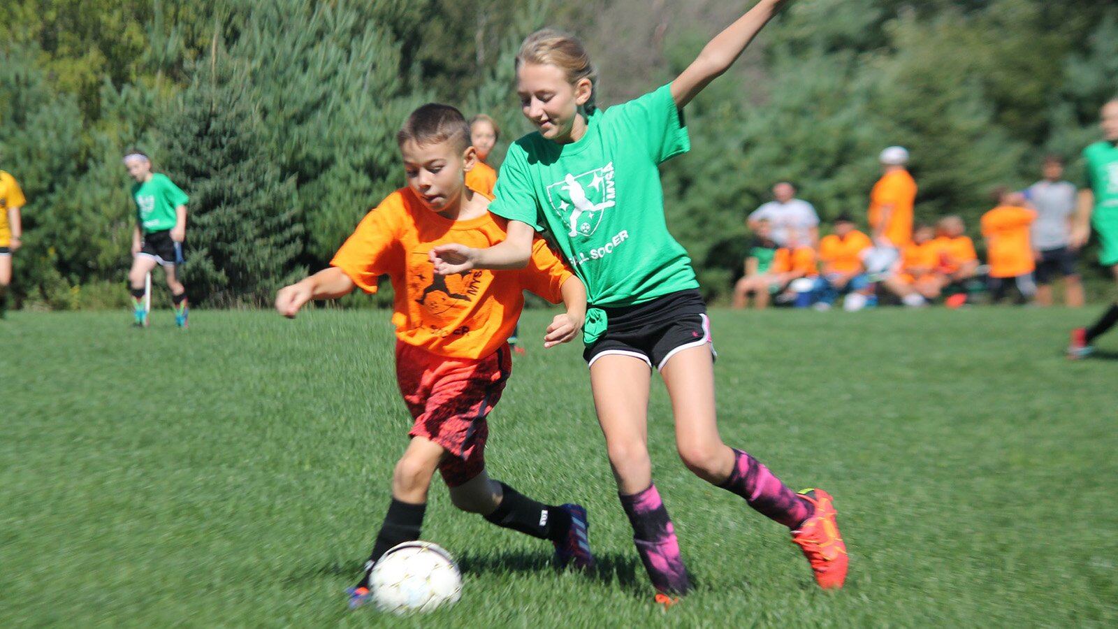 Zdjęcie przedstawia chłopca i dziewczynkę walczących o piłkę na murawie. Chłopiec ubrany jest w strój piłkarza – pomarańczową koszulkę i czerwone szorty, zaś dziewczynka w zieloną koszulkę i czarne szorty. W tle widoczna jest grupa osób w pomarańczowych koszulkach.