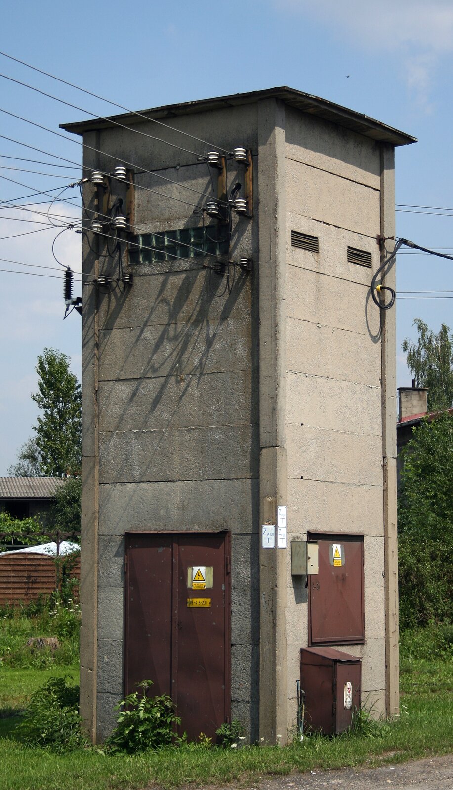 Rys. 4. to zdjęcie wieży transformatorowej w Woli Hankowskiej. Wieża, widoczna na tle niebieskiego nieba i zielonej roślinności i fragmentów zabudowań, ma postać pionowego cementowego prostopadłościanu widzianego z boku. Na dole wieży zamontowano metalowe drzwi w kolorze brązowym oraz na sąsiedniej ścianie - drzwiczki metalowe na wysokości połowy wysokości drzwi oraz obok - skrzynia metalowa, najprawdopodobniej na piasek. W górnej części ściany zamontowano konstrukcję utrzymującą układ transformatorów od których odchodzą linie elektryczne.