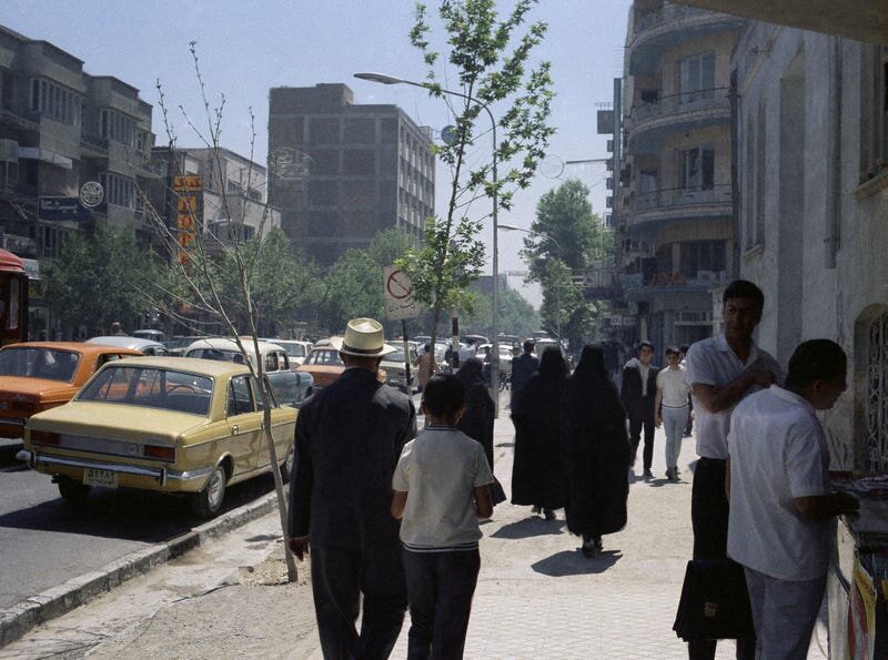 Zdjęcie przedstawia ludzi na chodniku. Wysoka zabudowa  miejska. Zatłoczone ulice. Kobiety w chustach na głowie. 