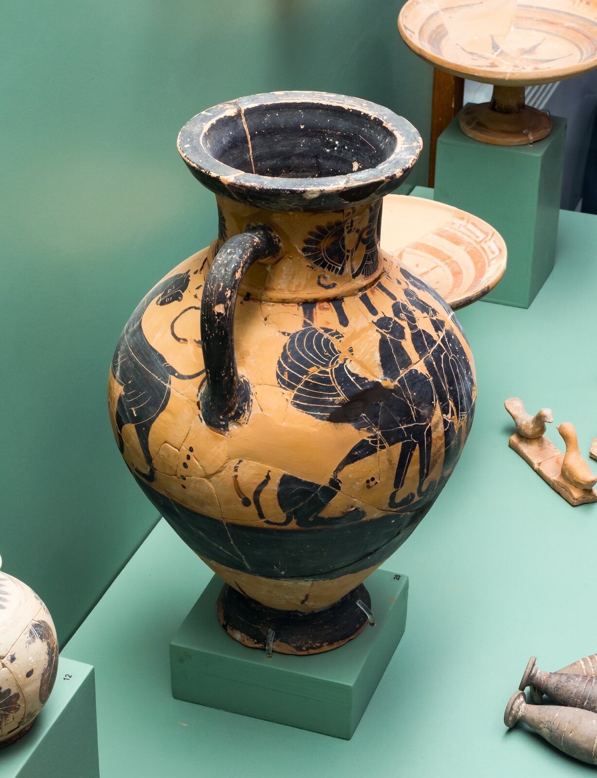 Fotografia autorstwa ArchaiOptix przedstawia starożytną amforę, na której namalowana została tak zwana Grupa z Rodos - jest to grupa osób (kobiet i mężczyzn) stojąca obok mitologicznych stworzeń - wyróżnić można tutaj sfinksa. Naczynie zostało wykonane w stylu czarnofigurowym. Obok widoczne są inne naczynia, kształtem przypominające płytkie talerze.