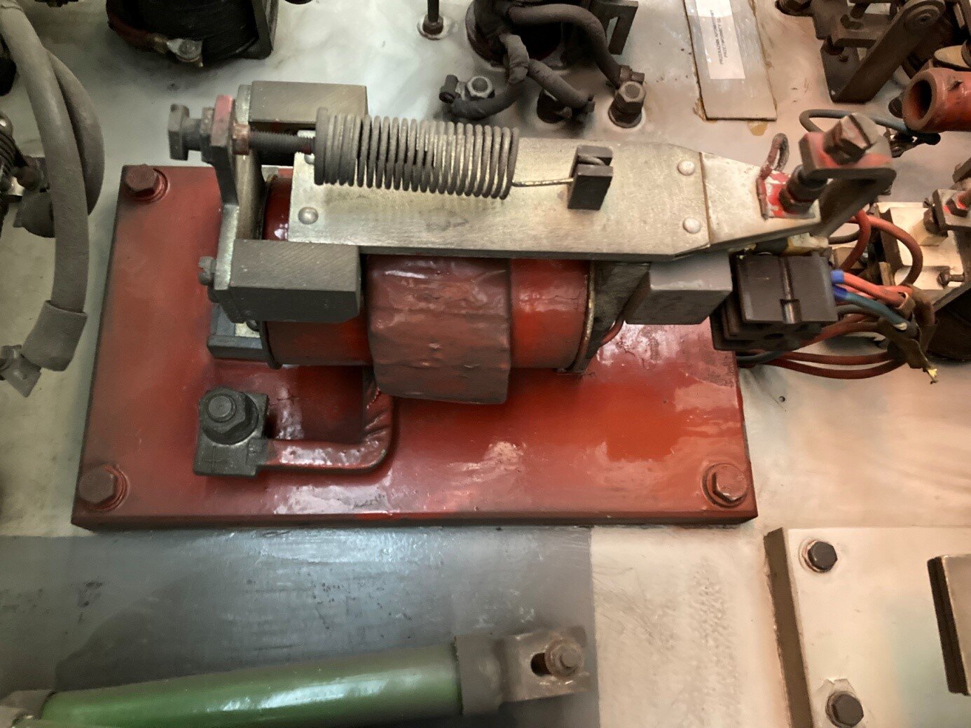 Na zdjęciu widoczny jest przekaźnik samoczynnego rozruchu. Przekaźnik umieszczony jest na czerwonej podstawie przykręconej do podłoża. Na podstawie przytwierdzony jest stelaż z poziomo umieszczonym walcem cewki pomocniczej otoczonym cewką prądową. Nad walcem na stelażu jest płyta zwana kotwicą z trójkątnym zakończeniem z prawej strony. Z kotwicy wystaje uchwyt, do którego podczepiona jest sprężyna odciągowa przykręcona do stelaża. Na trójkątnym zakończeniu kotwicy znajduje się styk sterujący wałem kułakowym. Odchodzi od niego przewód elektryczny.