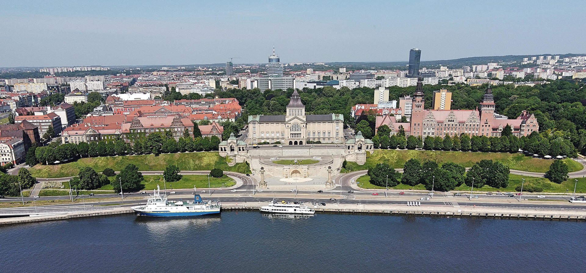 Zdjęcie lotnicze Szczecina, widok na Wały Chrobrego. W tle panorama miasta, w centralnej części duży budynek a przed nim betonowy plac. Na pierwszym planie Odra, zacumowane niewielkie statki. Wzdłuż brzegu droga.
