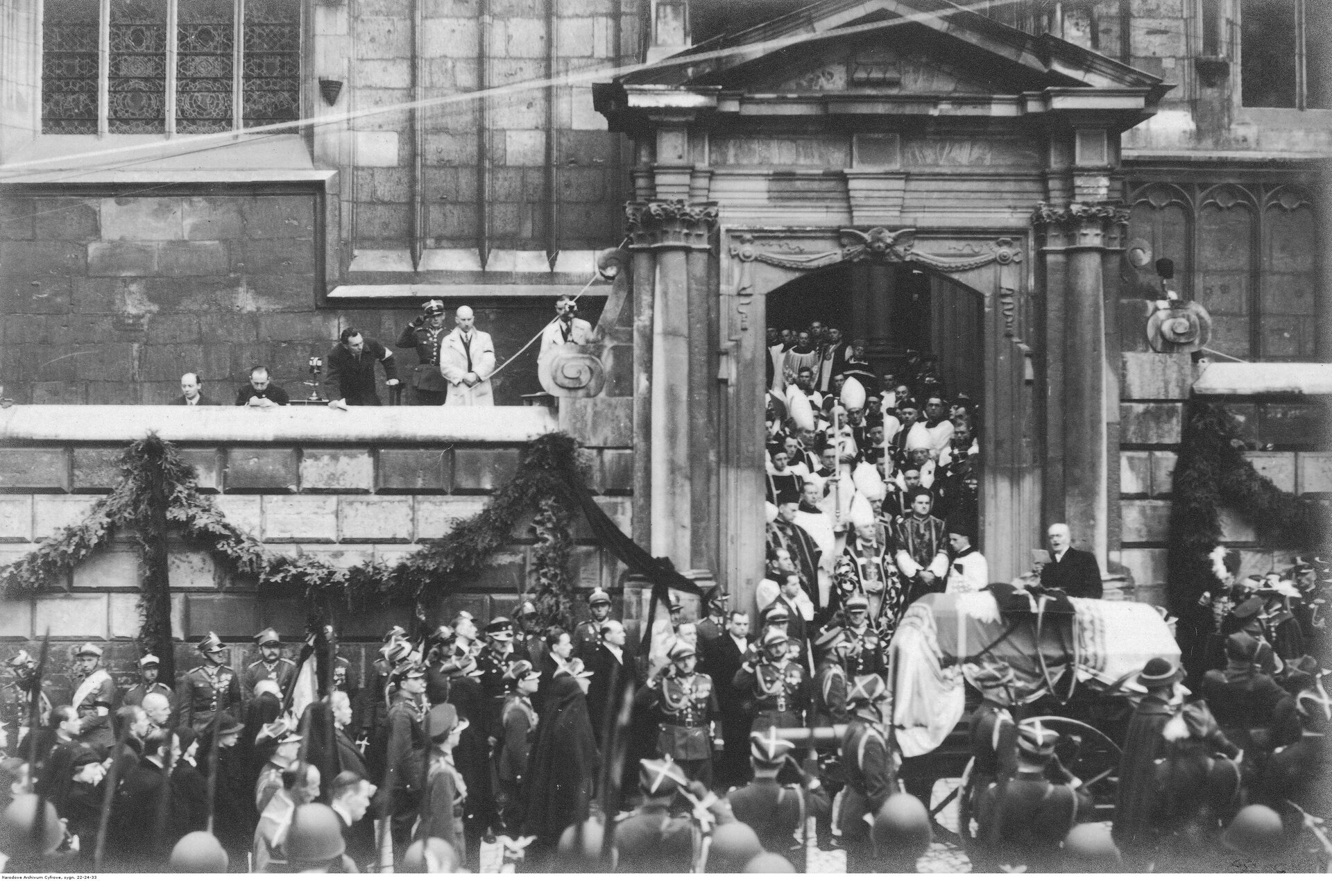 Zdjęcie przedstawia kondukt żałobny. Wychodzi on z Katedry na Wawelu. Tłum ludzi otacza trumnę z ciałem Józefa Piłsudskiego. Trumna jest na wozie. Jest przykryta sztandarem. Wśród tłumu są licznie zgromadzeni biskupi, księża i wojskowi. W kondukcie, tuż za trumną, widać najbliższą rodzinę marszałka.