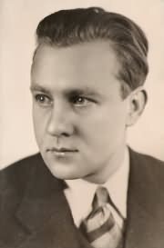 Na zdjęciu widać mężczyznę w garniturze i krawacie, ma krótkie, gładko zaczesane włosy. Tło jest jasne i neutralne