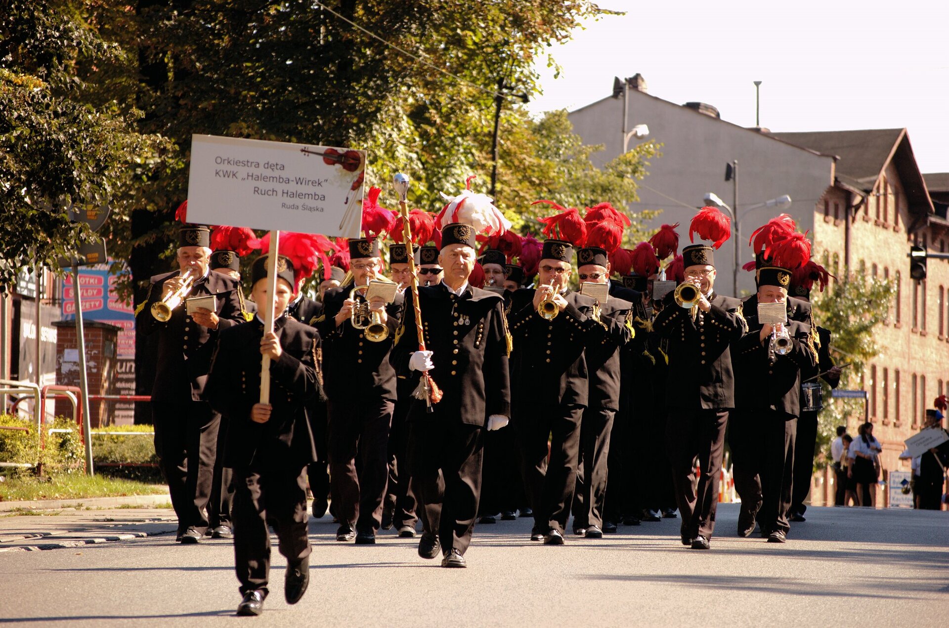 Fotografia kilkunastoosobowej orkiestry górniczej na ulicach miasta. Mężczyźni ubrani w czarne górnicze mundury, grają na instrumentach dętych. Noszą okrągłe, wysokie czapki z pióropuszami, zazwyczaj czerwonego koloru (jeden ma kolor biały). Przed nimi idzie chłopiec, również w górniczym stroju z planszą o treści: Orkiestra dęta KWK Halemba‑Wirek, Ruch Halemba, Ruda Śląska.
