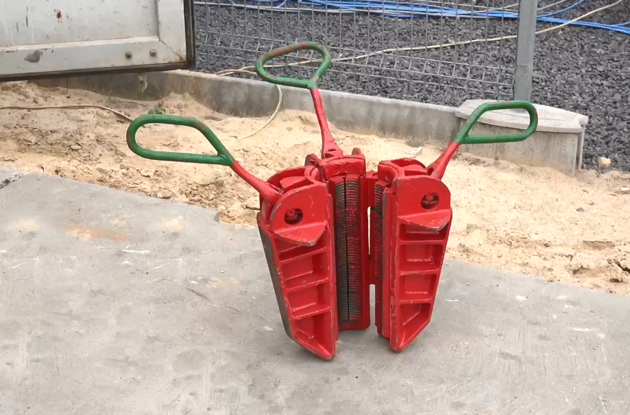 Na zdjęciu widoczny jest czerwony klin wiertniczy. Klin jest to narzędzie stosowane przy zapuszczaniu kolumn rur okładzinowych. Klin wykonany jest z trzech metalowych elementów - zębów. Elementy na zdjęciu są rozwarte. Na zewnętrznej powierzchni zęba zębów znajdują się nacięcia. W  górnej części klina znajduje się metalowy pręt zakończony okrągłą rączką.Widoczny klin położony jest na betonowej płycie.