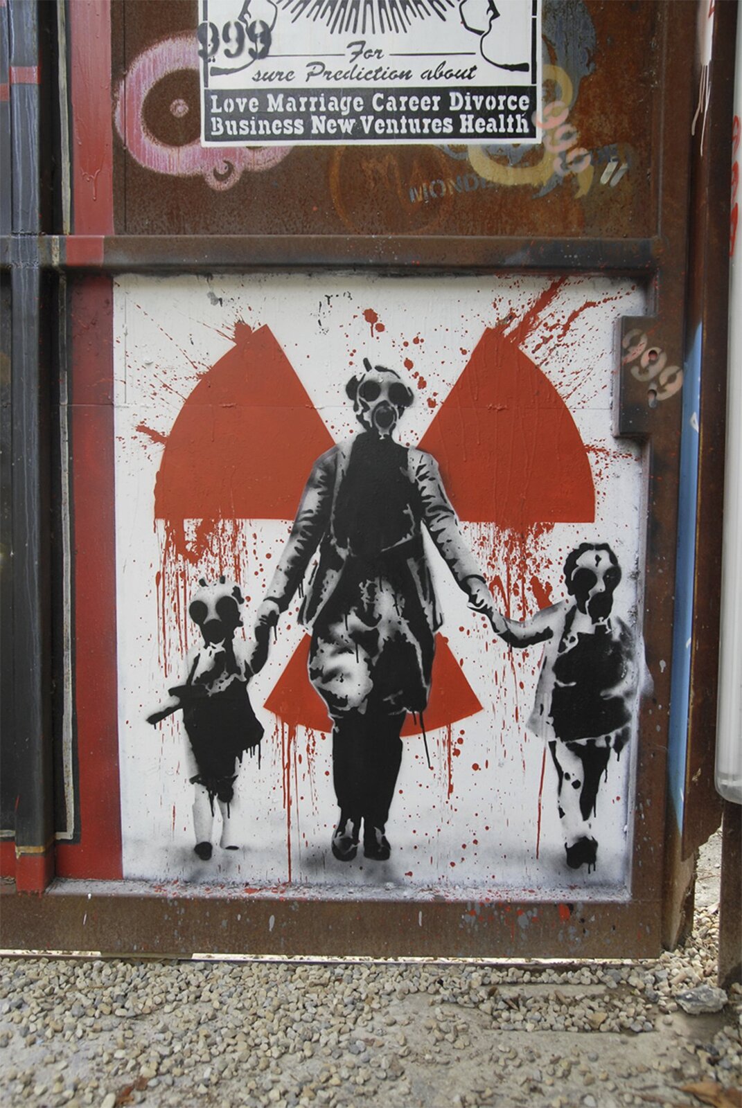 Zdjęcie przedstawia krytyczne w stosunku do energetyki jądrowej graffiti na stalowym kontenerze. Na białym tle namalowany jest czerwoną farbą wiatraczek, symbol ostrzegawczy przed promieniowaniem radioaktywnym. Na tle tego znaku znajdują się czarnobiałe postacie kobiety trzymającą dwójkę dzieci za dłonie. Cała trójka ma na twarzach maski przeciwgazowe.
