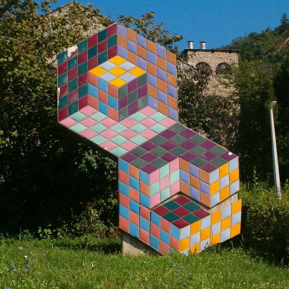 Ilustracja interaktywna przedstawia wolno stojącą rzeźbę Victora Vasarely. Rzeźba wykonana jest z porcelany. Kształtem przypomina pochyloną cyfrę osiem. Składa się z małych, kolorowych rombów. Dzieło znajduje się na trawniku. W tle widoczne są liczne drzewa oraz fragment budowli. Zestawienie ze sobą romboidalnych kształtów oraz barw dopełniających daje efekt iluzji trójwymiarowości. Ulegamy złudzeniu głębi, widząc w figurze dwie prostopadłościenne przestrzenie, w których znajdują się sześciany.