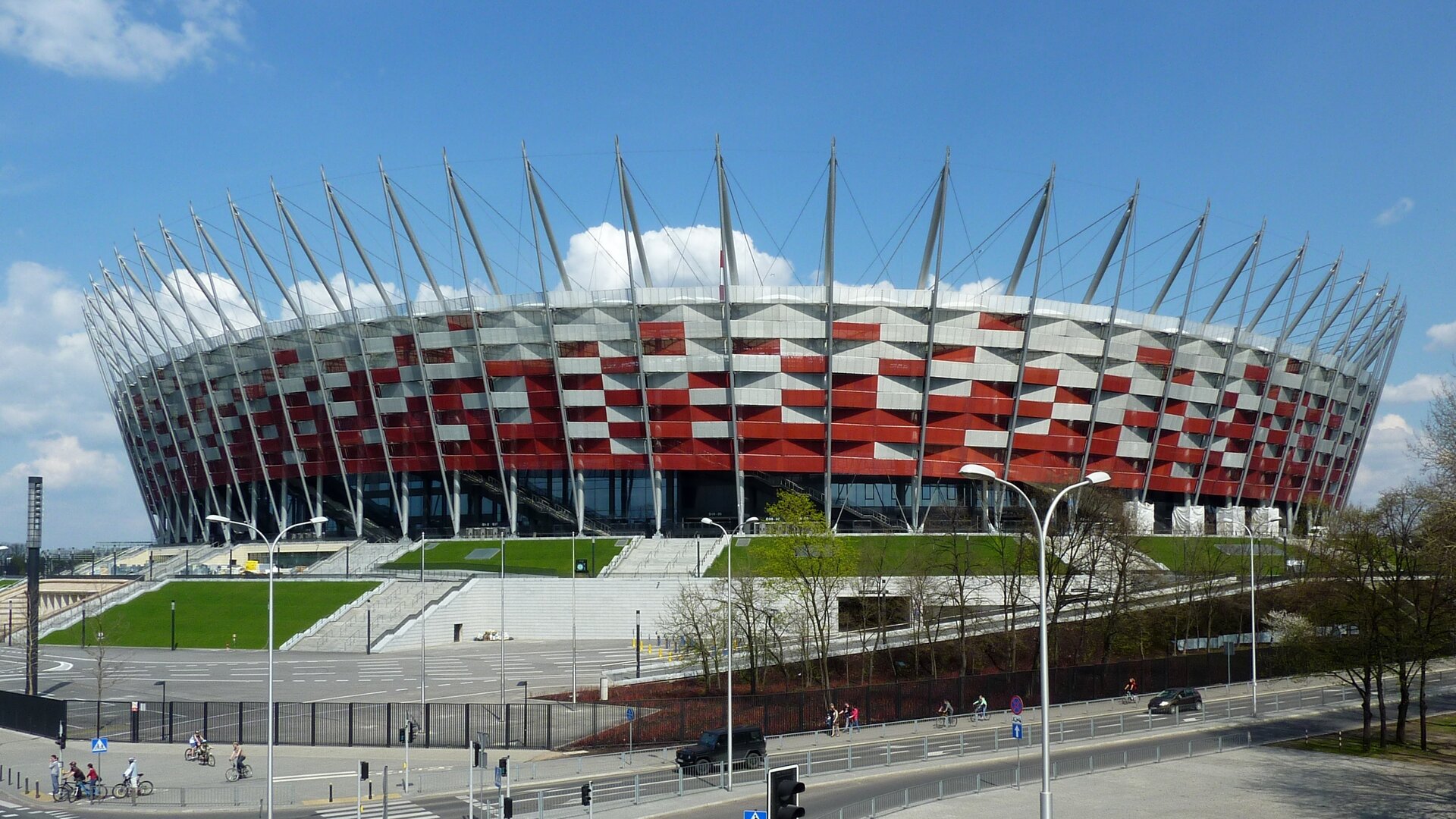 Fotografia przedstawia widok na Stadion Narodowy w Warszawie.  Jest to duży budynek z biało‑czerwoną fasadą, na której przeplatające się prostokąty koloru czerwonego i białego przypominają swoją strukturą wiklinowy kosz. Ściana budynku jest zaokrąglona, u jego podstawy widoczne są duże szyby. Można również zauważyć biegnące od dołu do góry, a także do wewnętrznej strony budynku grube, srebrne belki, tworzące jego konstrukcję. Przed nim widoczne są schody, a także fragmenty zielonego terenu oraz ulicę przebiegającą obok. Fotografia została wykonana w lekko zachmurzony dzień.