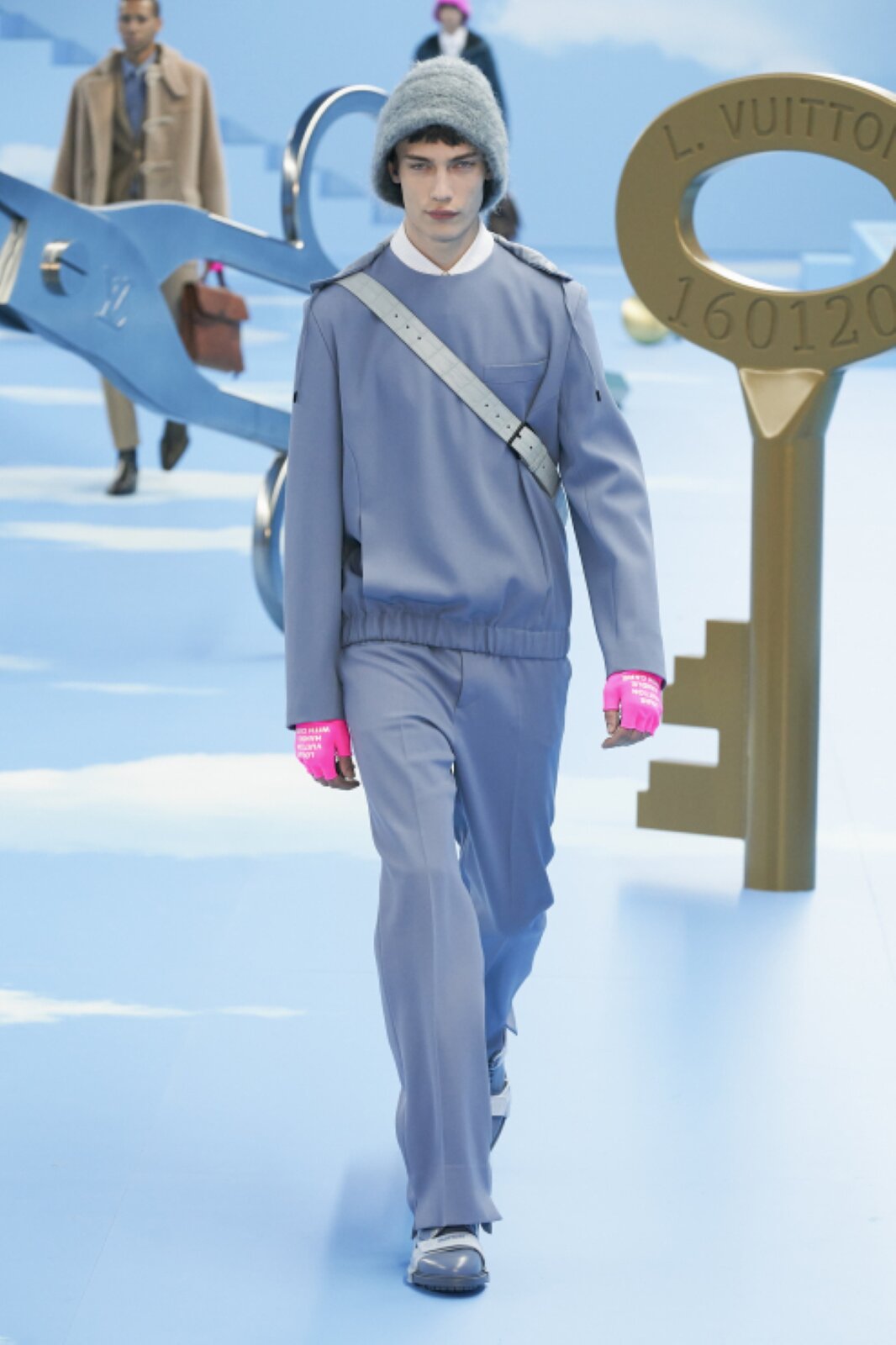 Fotografia przedstawia ciemnowłosego mężczyznę, który ma na sobie komplet składający się z luźnych spodni zaprasowanych na kant oraz luźnej bluzy w tym samym jasnym odcieniu koloru niebieskiego. Przez jego tors przechodzi pas niewidocznej torby, prawdopodobnie spoczywającej na jego plecach. Na głowie mężczyzny widoczna jest jasna czapka w stylu „bucket hat”, a na jego dłoniach jaskrawe różowe rękawiczki odsłaniające palce z białymi napisami.