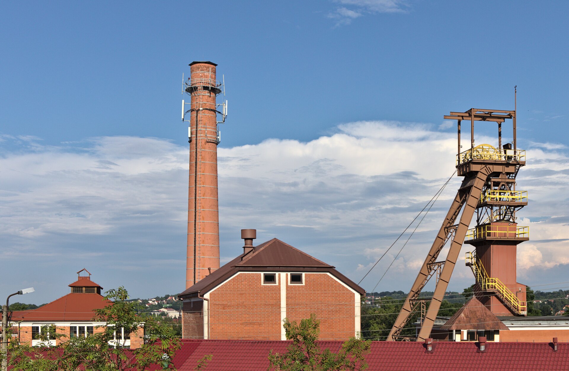 Zdjęcie ukazuje budynki kopalni soli w Bochni. Widoczny jest budynek z pomarańczowej cegły, za nim stoi komin, obok budynku znajduje się szyb.