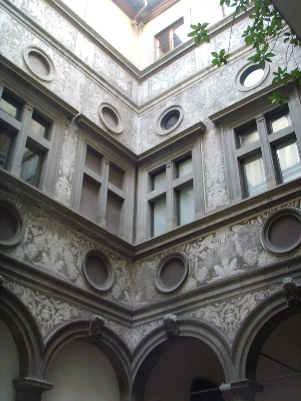 Widok w górę na wewnętrzny dziedziniec renesansowego pałacu. Ściany wyłożone są jasnym tynkiem pokrytym dekoracyjnymi sgraffitami o motywach roślinnych i figuralnych. Kondygnacje rozdzielają wyraźne gzymsy, a w pasie międzyokiennym rozmieszczone są okrągłe oculi. Prostokątne okna ujęte są w kamienne obramienia. Na dole widoczne są arkady wsparte na kolumnach z kapitelami, tworzące krużganek wokół dziedzińca. W prawym górnym rogu kadru widać fragment zielonych liści zwisających z góry.