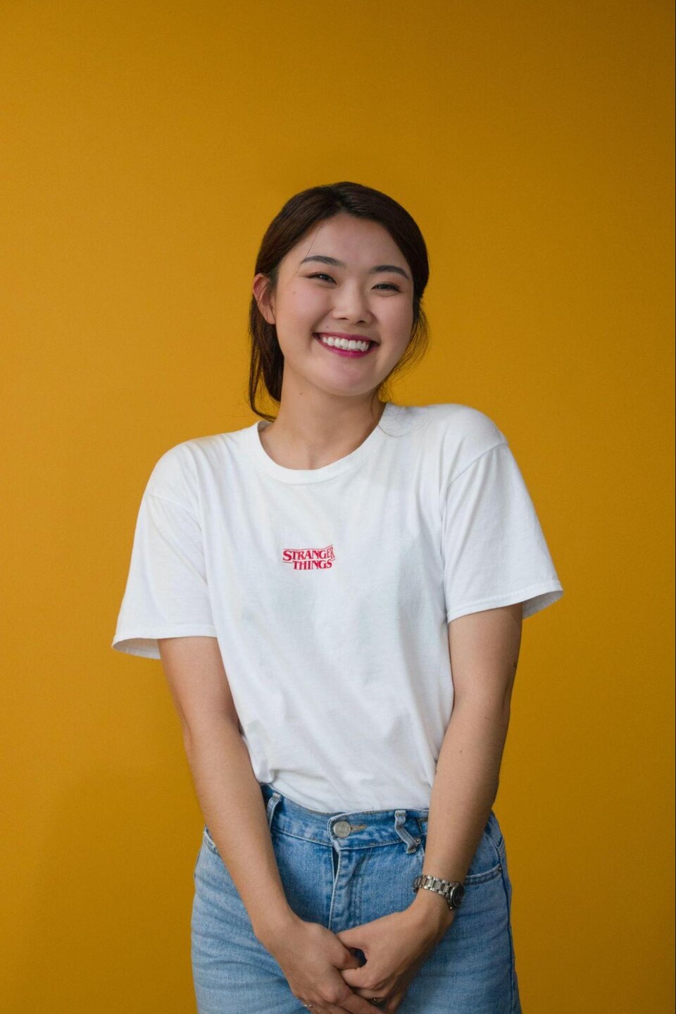 Zdjęcie przedstawia młodą dziewczynę ubraną w białą koszulkę i dżinsy. Ma azjatycką urodę. Tło zdjęcia jest żółte. 