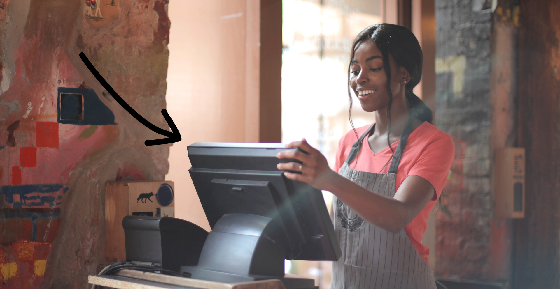 Zdjęcie przedstawia uśmiechniętą, czarnoskórą dziewczynę stojącą przed ekranem kasy. Strzałka skierowana jest na kasę.