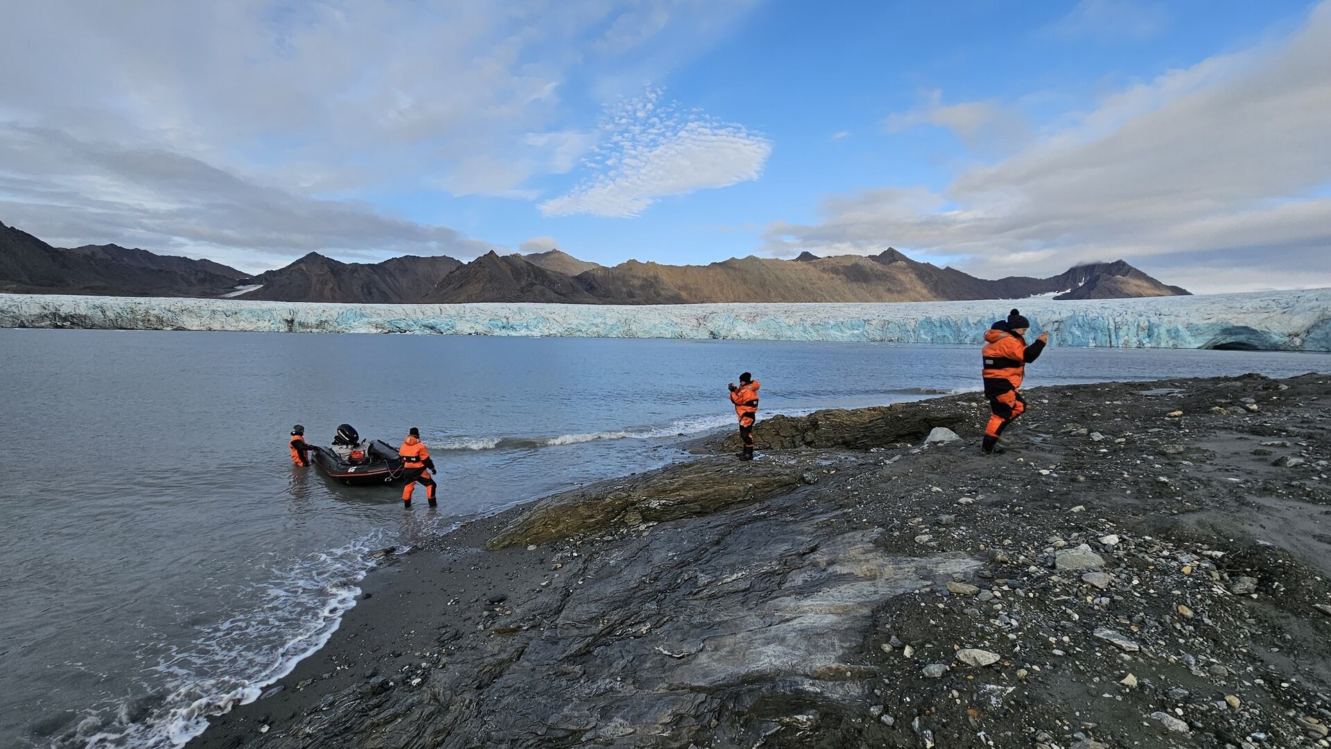 Fotografia kilku osób w czarno‑pomarańczowych grubych kombinezonach i czapkach nad brzegiem zbiornika wodnego. Dwójka z nich wyprowadza na skalisty brzeg ponton z silnikiem. Na drugim planie na wodzie unosi się ogromna masa lodu, na drugim planie pasmo rozległych gór.