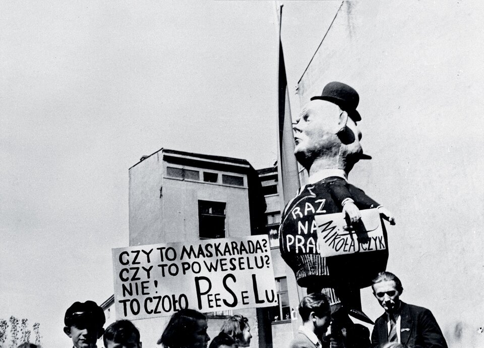 Zdjęcie przedstawia grupę ludzi na ulicy. Widać trzymaną przez nich kukłę przedstawiającą Stanisława Mikołajczyka, a także transparent z napisem "Czy to maskarada? Czy to po weselu? Nie! To czoło PeeSeLu.