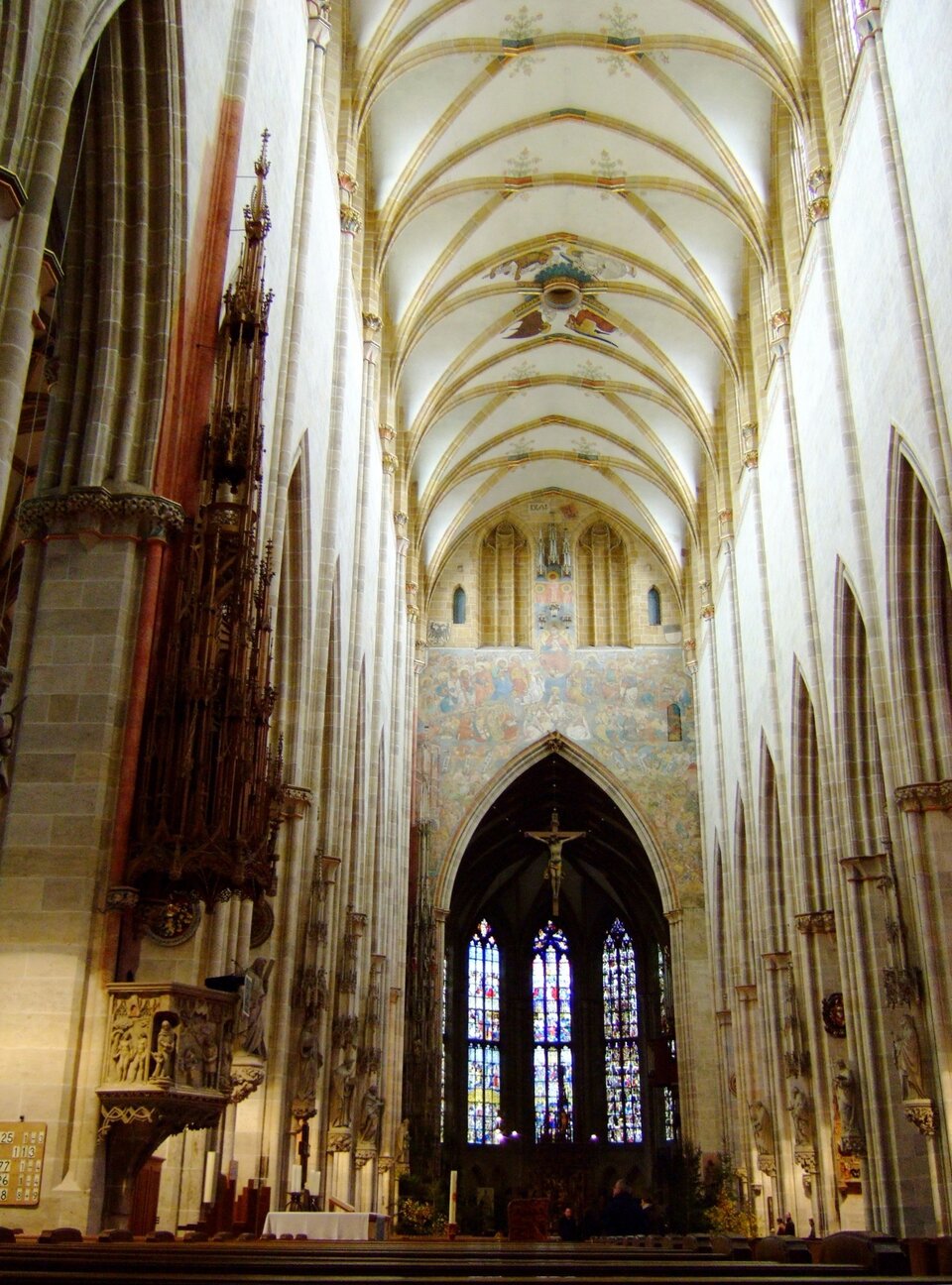 Wnętrze katedry Najświętszej Marii Panny w Ulm; widok nawy głównej, na dalszym planie zawieszony wysoko krzyż, za nim trzy wysokie okna witrażowe.
