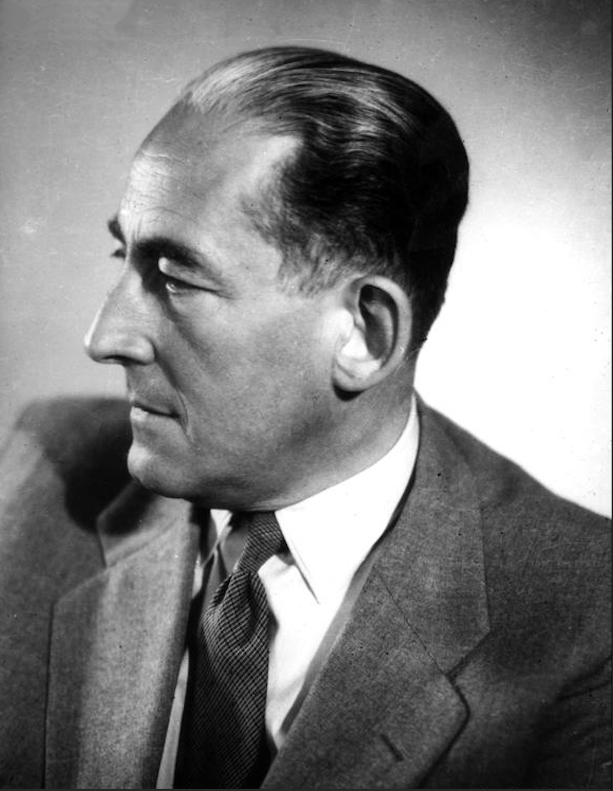 Czarnobiałe zdjęcie przedstawia portret mężczyzny w średnim wieku. Postać jest widoczna z profilu. Ma ciemne, lekko siwiejące włosy. Jest ubrana w biała koszulę z kołnierzykiem, krawat w drobny wzór geometryczny i szarą marynarkę. Tło jest jednolite. 