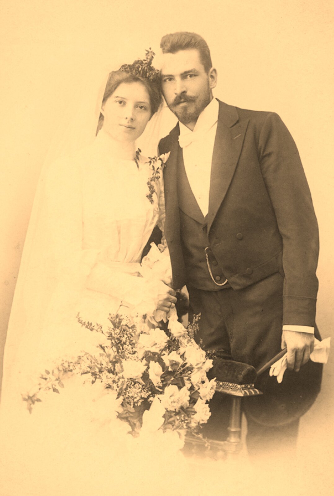 Rys. 4. Zdjęcie w sepii przedstawia ślubny portret Zofii i Mariana Smoluchowskich. Kobieta ma długą, białą suknię, biały welon upięty na włosach zaczesanych w skomplikowaną fryzuję. Mężczyzna ma ciemne włosy zaczesane do tyłu, przystrzyżoną brodę i wąsy. Ubrany jest w ciemny smoking, kamizelkę oraz białą koszulę z białą muszką pod szyją. W lewej ręce trzyma kapelusz i białe rękawiczki. Przed parą małżonków stoi duży bukiet jasnych kwiatów.