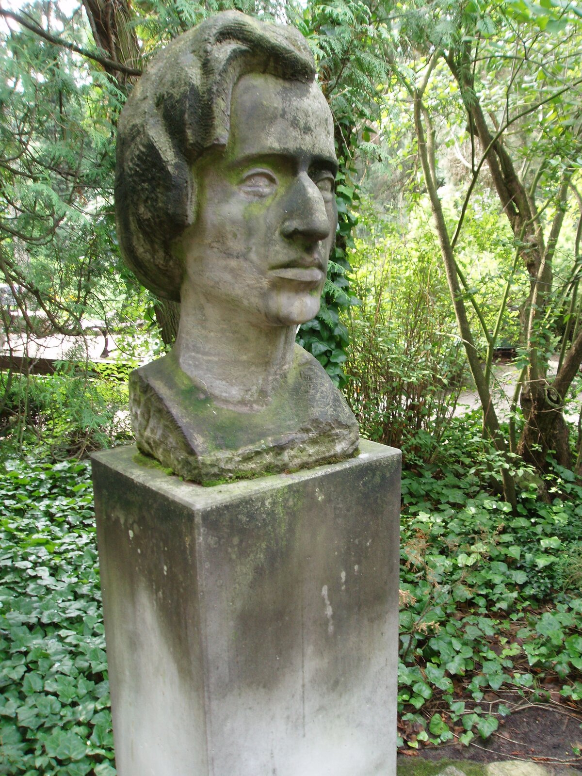 Zdjęcie przedstawia kamienne popiersie mężczyzny, ustawione na masywnym cokole. Mężczyzna ma duży, wystający nos oraz dość długie włosy. W tle znajduje się park.