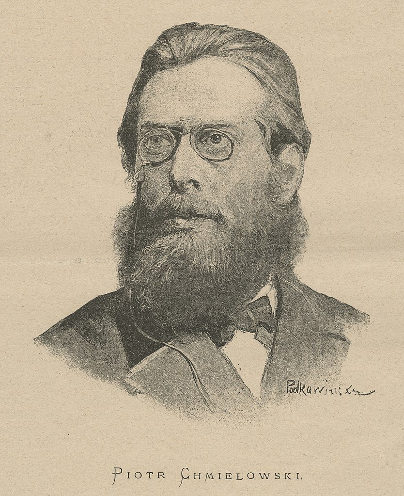 Ilustracja przedstawia mężczyznę w średnim wieku. Ma dłuższe włosy zaczesane do tyłu. Na nosie małe okulary z łańcuszkiem. Długą brodę oraz wąsy. Ubrany jest w elegancki garnitur oraz muszkę.