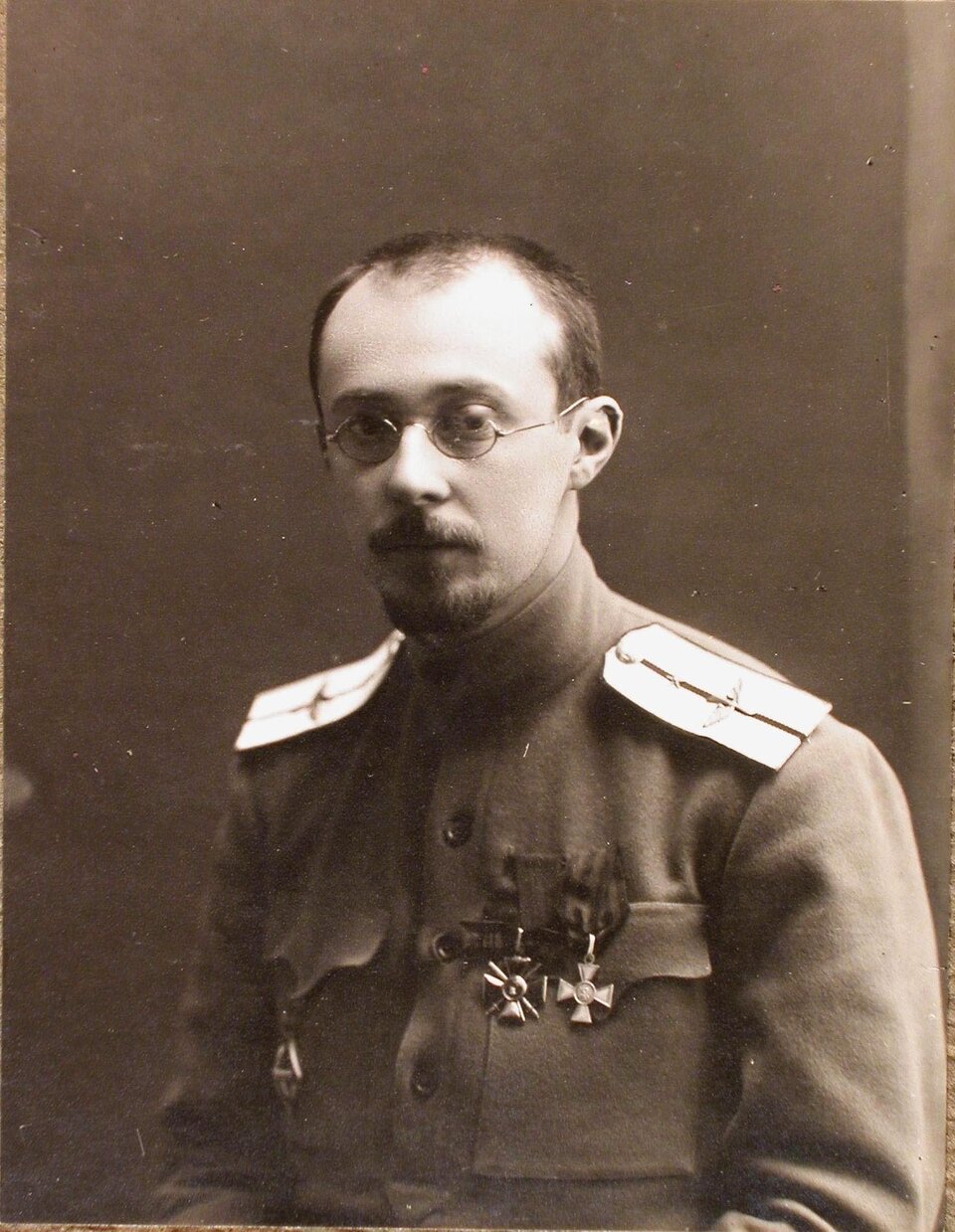 Zdjęcie portretowe przedstawia młodego mężczyznę mundurze wojskowym. Mężczyzna ma wysokie czoło,  zakola; nosi małe okrągłe okulary oraz krótką brodę. Na mundurze po lewej stronie ma przypięte dwa ordery.  