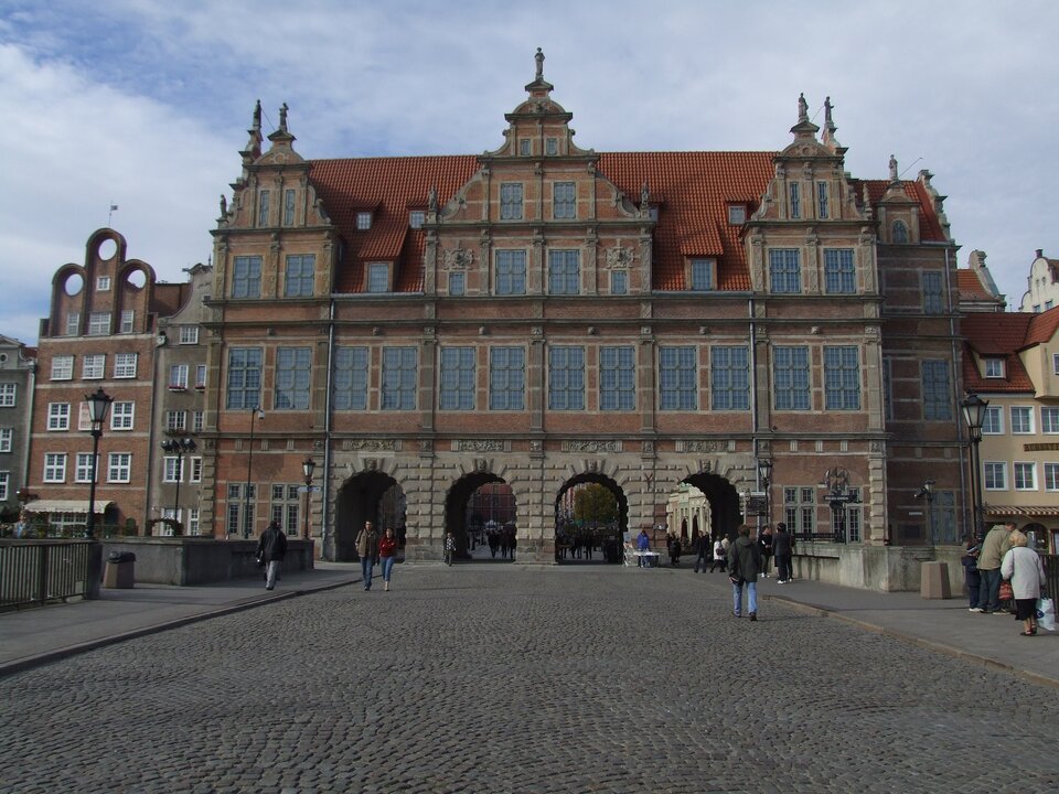 Zdjęcie przedstawia zabytkowy obiekt w Gdańsku od strony Długiego Pobrzeża, zwany Bramą Zieloną. Budynek utrzymany w stylu manieryzmu niderlandzkiego. Cztery otwory pełnołukowe prowadzą przez bramę na Długi Targ. Powyżej poziomu parterowego, oddzielonego gzymsem, rząd prostokątnych otworów okiennych. Potrójny szczyt, z dwoma rzędami okien każdy na bokach i szczyt z trzema rzędami pośrodku, osłania dwuspadowy dach, pomiędzy szczytami lukarny, Na szczytach figury postaci męskich i żeńskich. Budynek wykonany z cegły i kamienia. Do bramy prowadzi bruk położony na Zielonym Moście. Na moście spacerują przechodnie.