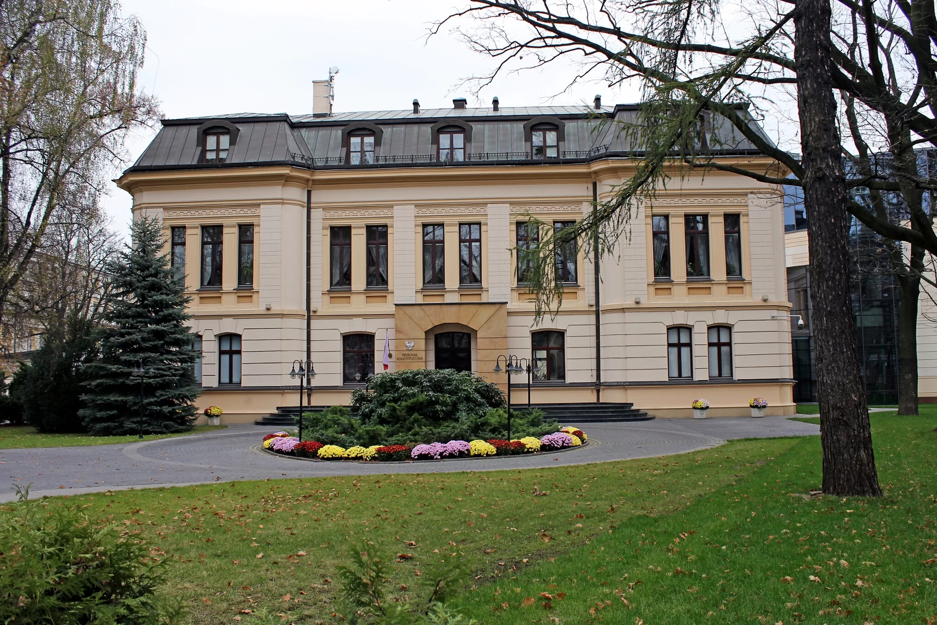 Kliknij żeby powiększyć Zdjęcie przedstawia dwukondygnacyjny budynek zwieńczony mansardowym dachem z oknami. W pierwszej kondygnacji budynek ozdobiony jest boniowaniem. W centrum w części wysuniętej nieznacznie przed bryłę budynku znajduje się wejście do którego prowadzą schody. Wysokie okna w pierwszej kondygnacji oddzielone są od siebie pilastrami. Nad i pod nimi są elementy dekoracyjne. Przed budynkiem jest dziedziniec wyłożony kostką i okrągły klomb z kolorowymi kwiatami i zielonymi krzewami.