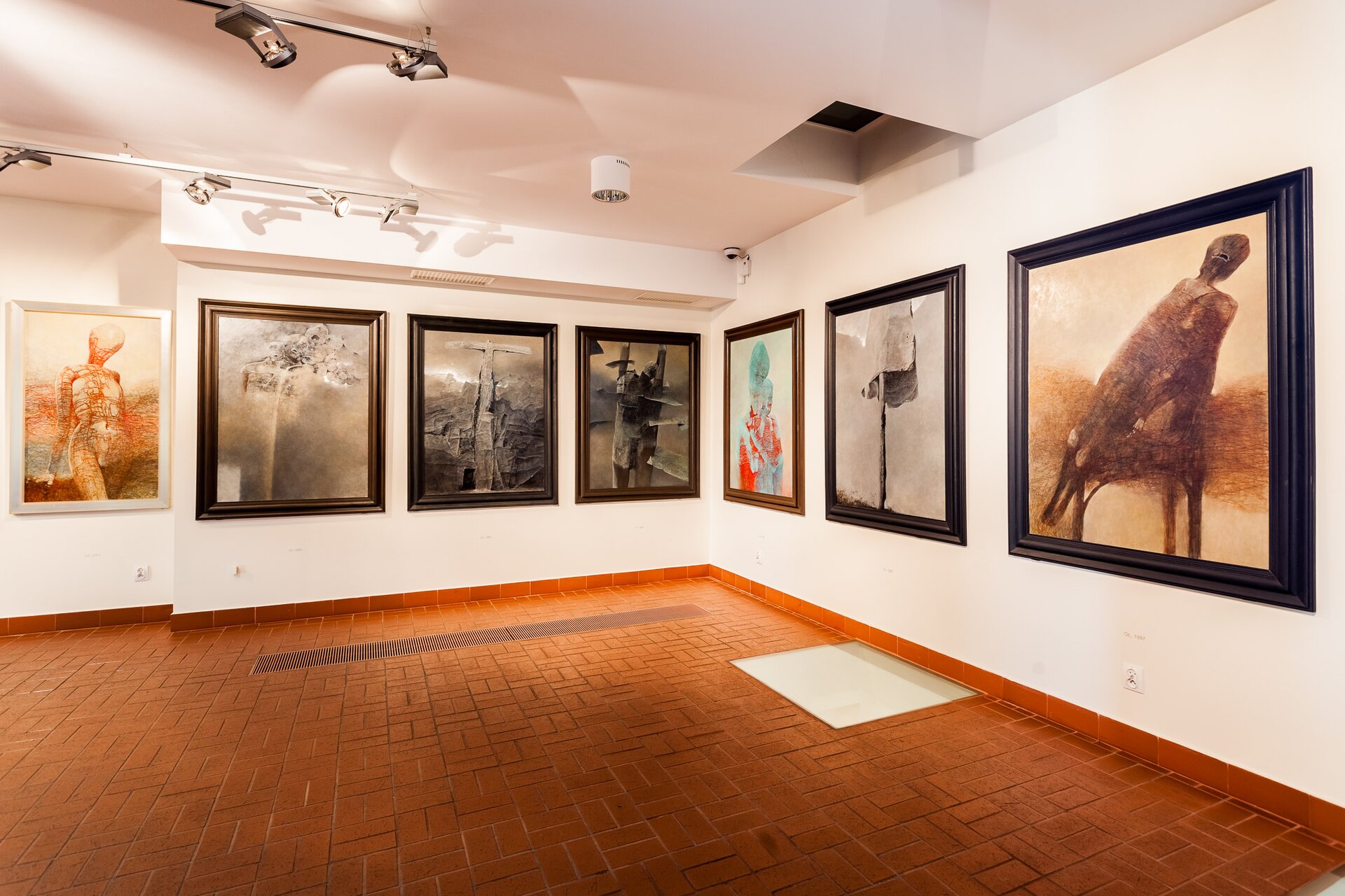 Zdjęcie przedstawia róg pomieszczenia w muzeum. Na białych ścianach wiszą ustawione symetrycznie obrazy w drewnianych ramach. Obrazy przedstawiają zarysy postaci podobnych do ludzkich, elementy w kształcie krzyża, zamazane, zamglone tło.