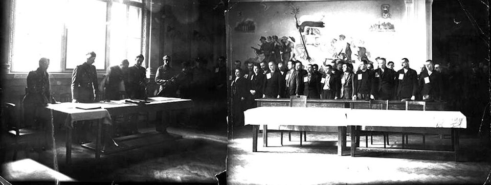 Zdjęcie przedstawia rozprawę odbywającą się przed sądem wojskowym. Widzimy ludzi zgromadzonych w sali o dużych oknach. Po lewej stronie, pod oknami stoi czterech mężczyzn w mundurach. Jeden z nich pochylając się nad rozłożonymi przed nim dokumentami odczytuje wyrok. Obok nich stoi żołnierz z karabinem. Po prawej stronie, na miejscu dla oskarżonych stoi kilkunastu mężczyzn. Ubrani są oni w stroje cywilne: marynarki, spodnie i koszule, a na prawej piersi mają przypięte kartki. Głowy mają zwrócone lekko w stronę sędziów. Przed nimi stoi długi stół i puste krzesła, co wskazywać może, że na sali nie było ich obrońców. Oskarżeni stoją na tle ściany, na której namalowani są walczący żołnierze i kobieta z biało‑czerwoną flagą  w dłoni.  