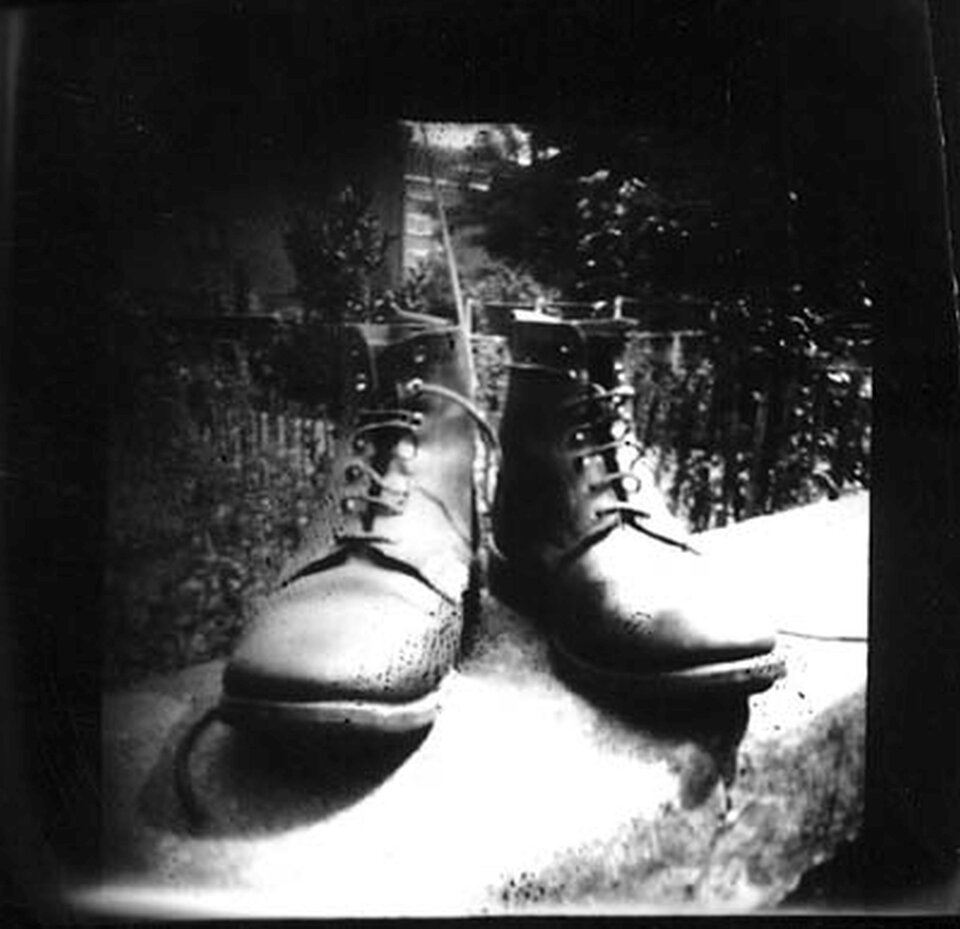 Ilustracja przedstawia zdjęcie Wolfa Howarda, „Buty”. Ukazuje stojące na murku skórzane buty z cholewkami. Stykają się piętami, a czubki rozchodzą się na zewnątrz. Fotografię wykonano techniką czarno-białą za pomocą camera obscura. Elementy znajdujące się bliżej przyrządu są nieostre. Murek oraz czubki butów są prześwietlone. W tle znajduje się pejzaż.