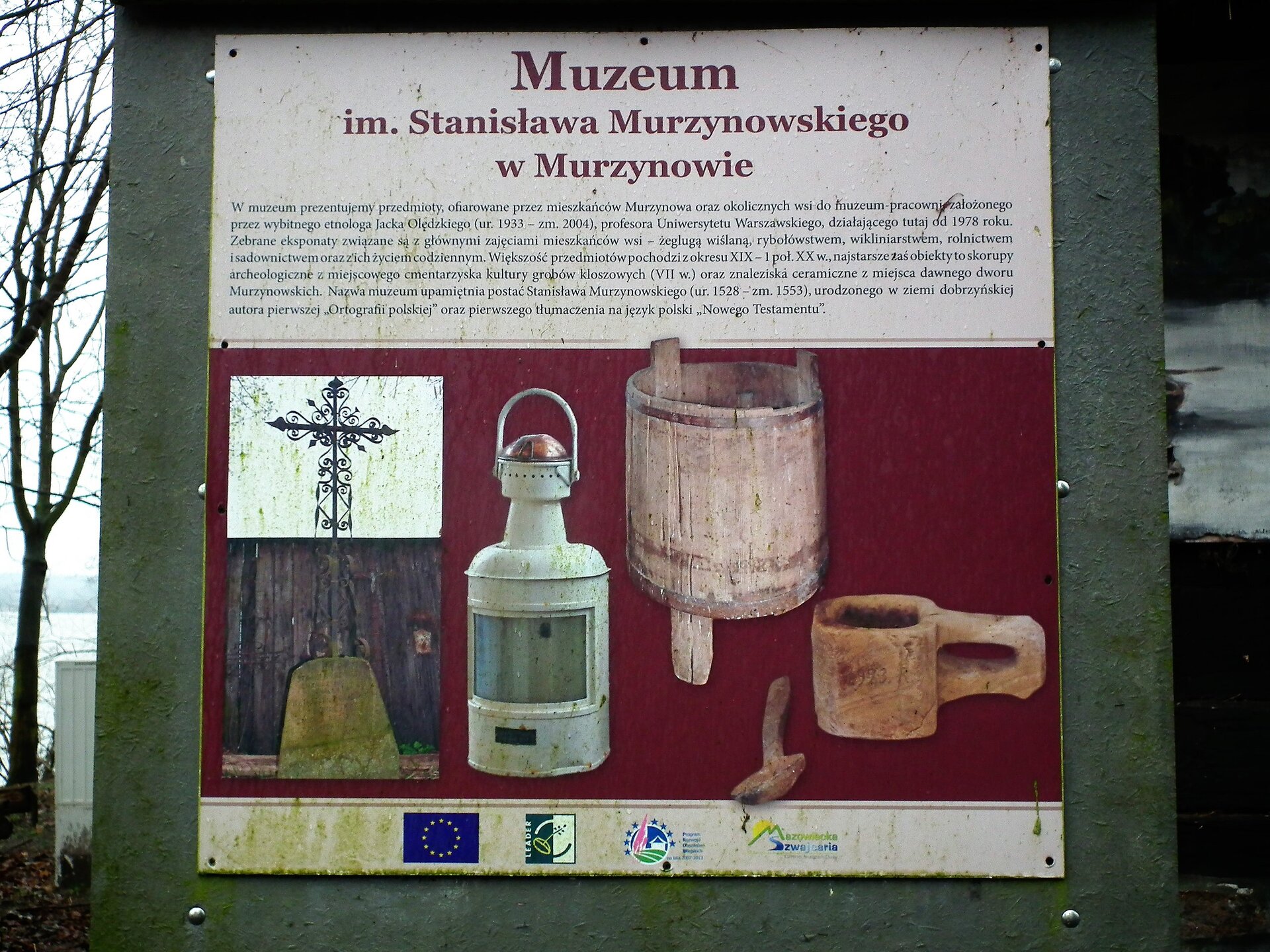 Fotografia przedstawia tablicę informacyjną u mieszoną przy muzeum im. Stanisława Murzynowskiego w Murzynowie. Na białym tle czerwone napisy. Pod spodem zdjęcia od lewej: krzyża, latarni, drewnianej beczki i glinianego kubka z łyżką. 