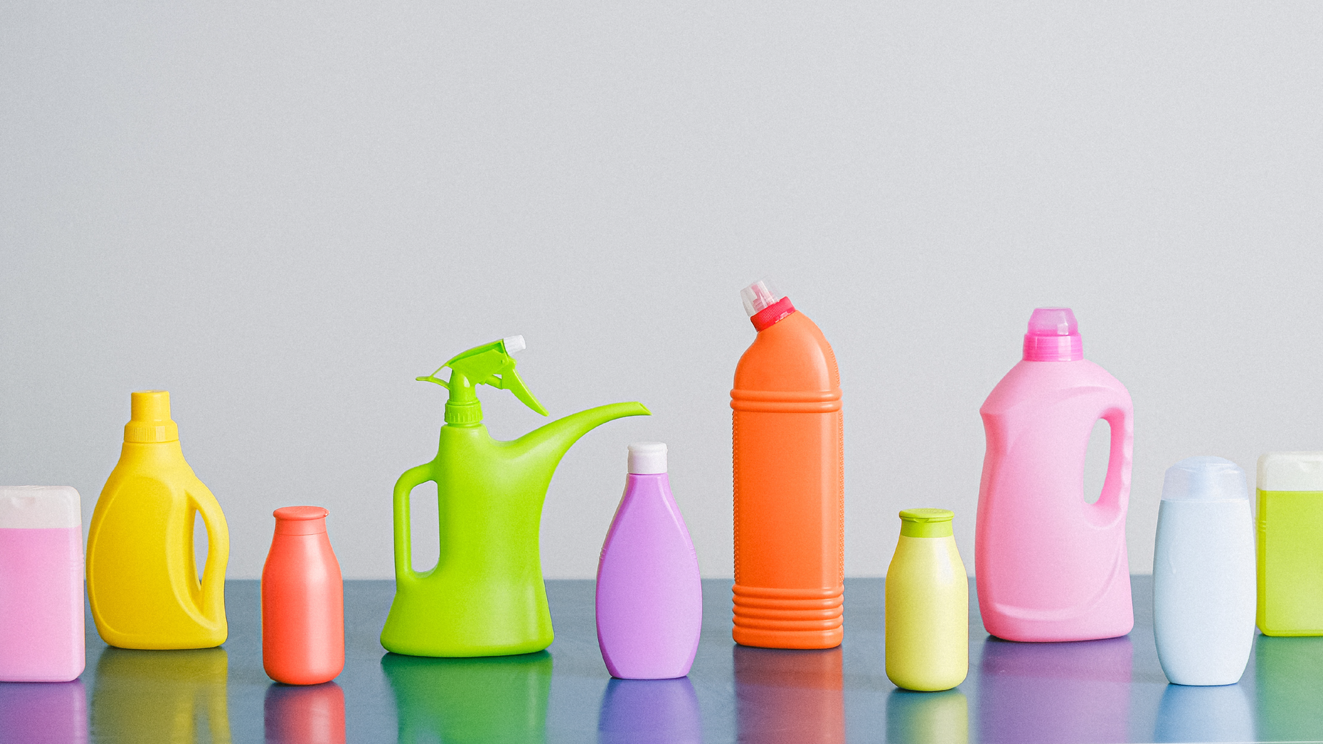 Zdjęcie przedstawia dziesięć pojemników ze środkami czystości. Pojemniki są ustawione na blacie w rzędzie, jeden obok drugiego. Pojemniki są różnej wielkości i w różnych kolorach. Na blacie stoją mniejsze i większe butelki. Nie ma na nich etykiet.