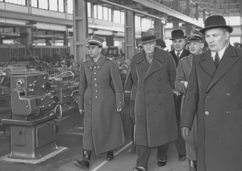 Zdjęcie przedstawia grupę mężczyzn w płaszczach, która idzie wzdłuż hali fabryki. Po lewej stronie znajdują się rzędy maszyn.