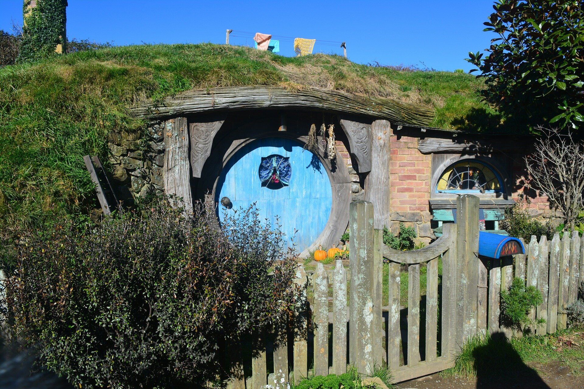 Kolorowe zdjęcie przedstawia domek hobbita wbudowany w pagórek porośnięty trawą. Przed domkiem znajduje się drewniany płotek z furtką. Na płocie wisi niebieska skrzynka na listy. Po obu stronach domku rosną drzewa i krzewy. Drzwi domku są okrągłe i niebieskie, po prawej stronie drzwi widać  półokrągłe okno. 