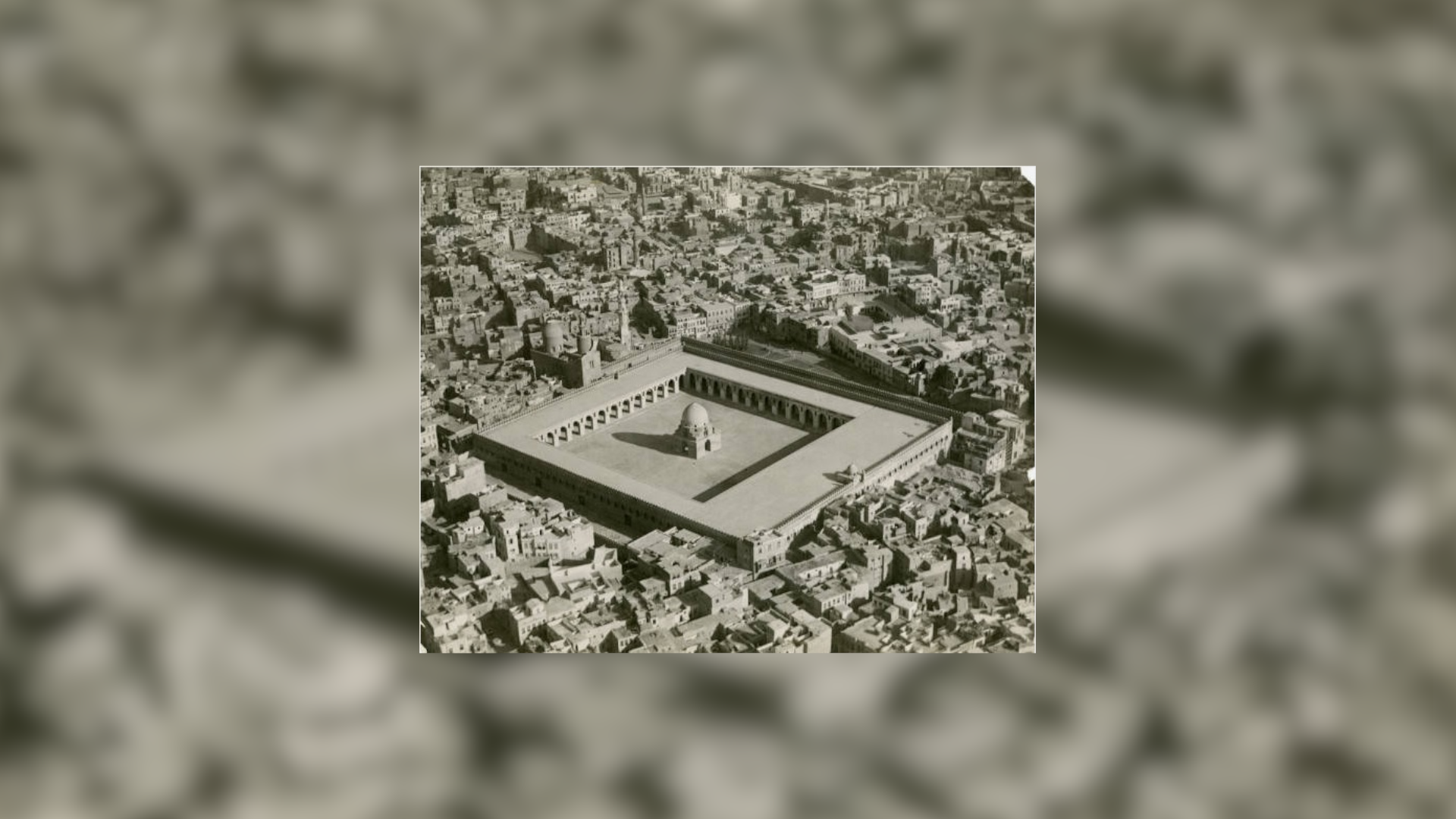 Czarno‑biała fotografia zrobiona z lotu ptaka przedstawia meczet znajdujący się w centrum miasta. Posiada on grube mory, duży plac ze świątynią na środku oraz minaret. 