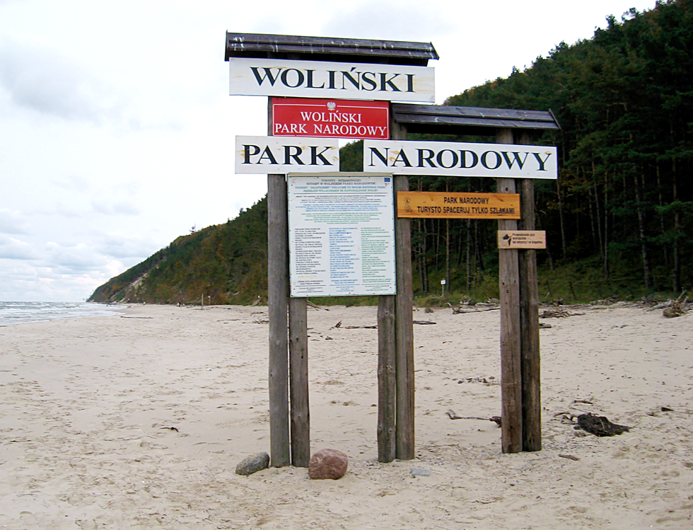 Fotografia przedstawia tablice informacyjne umieszczone na palach znajdujących się na plaży nadmorskiej Wolińskiego Parku Narodowego. Ilustracja przedstawia znak informujący o parku narodowym.