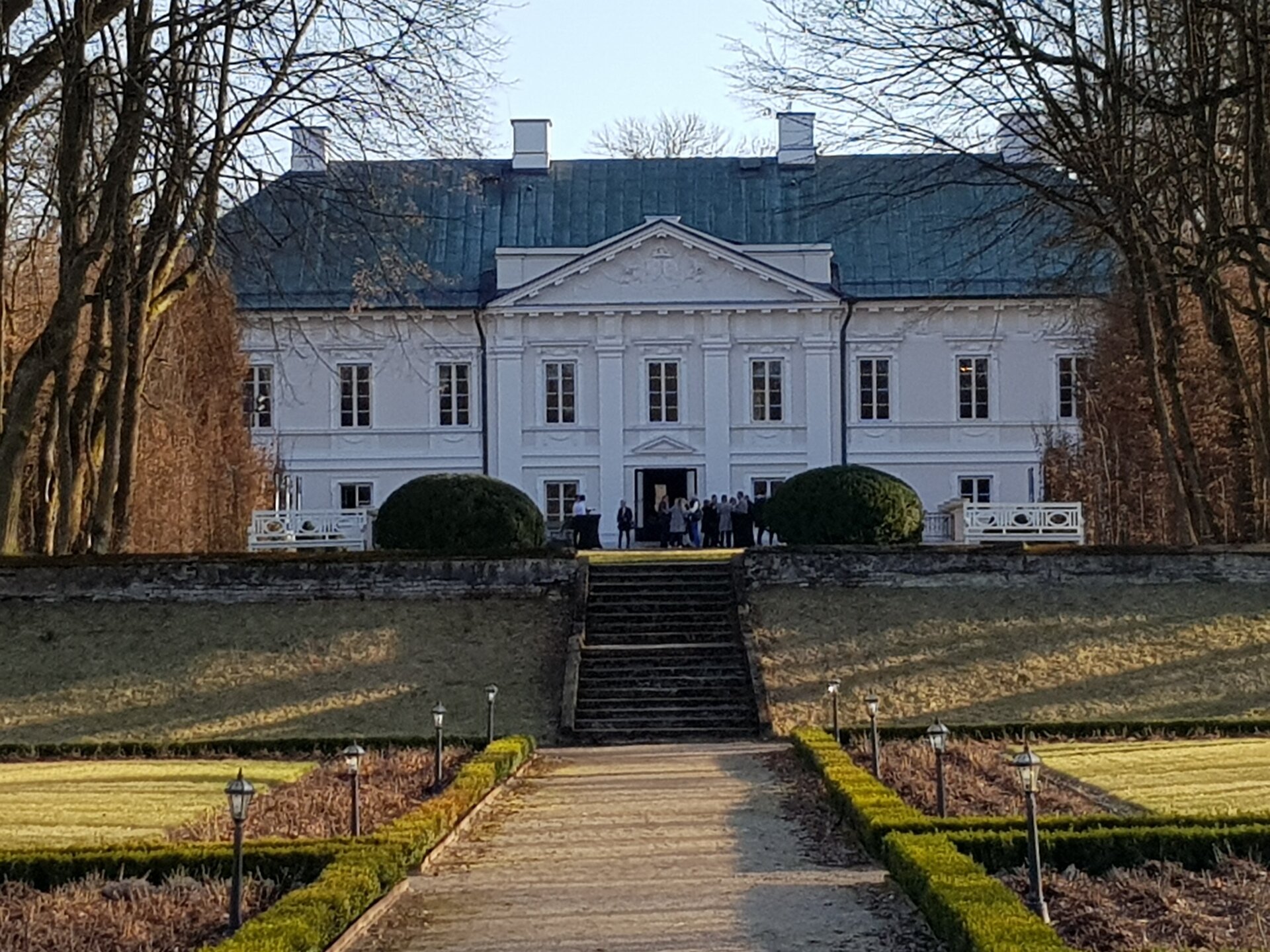 Zdjęcie przedstawia widok współczesny na pałac w Małej Wsi. Ścieżka przez ogród pałacowy prowadzi do szerokich schodów, następnie w kierunku pałacu. Centralna jego część to wysunięty fragment ściany z pilastrami i tympanonem, za którym znajduje się fragment attyyki, osłaniający czterospadowy dach. Na szczycie widoczne cztery kominy. Budynek utrzymany w stylu klasycystycznym, równo rozłożona linie okien na parterze i na piętrze. Przed drzwiami do wnętrza pałacu grupa turystów. Przed pałacem, powyżej schodów, dwa klomby, symetrycznie ustawione po bokach, przy nich ławki parkowe. Drogę do pałacu flankują lampy parkowe, wystające niewiele ponad linię niskiego żywopłotu. Drzewa na obu krawędziach fotografii bezlistne.