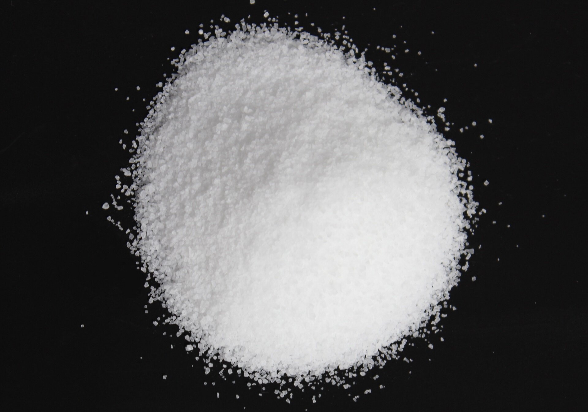 Na ilustracji znajduje się usypany na czarnej powierzchni, widziany z góry, kopczyk białej substancji, przypominającej cukier.