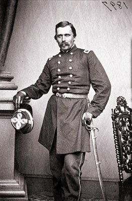 Zdjęcie przedstawia dojrzałego mężczyznę z wąsami i krótkimi włosami. Ubrany jest w mundur. W prawej ręce trzyma czapkę, a w lewej szablę.