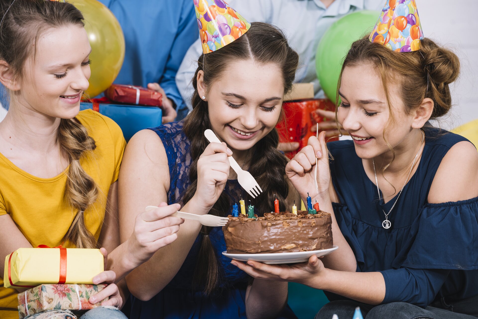 Zdjęcie przedstawia trzy uśmiechnięte nastolatki, które jedzą czekoladowy tort za pomocą jednorazowych widelczyków. Na torcie widoczne są różnokolorowe zdmuchnięte świeczki urodzinowe. Nastolatki mają na głowach kolorowe czapeczki urodzinowe, a jedna z nich trzyma na kolanach prezenty owinięte w kolorowe papiery i przewiązane wstążkami. W tle za nimi widoczne są fragmenty dwóch innych postaci, które trzymają na kolankach pudełka prezentowe.
