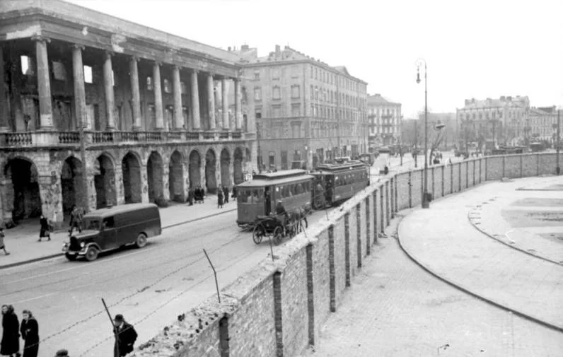 Czarno‑białe zdjęcie przedstawia mur getta. Jest on wysoki i zbudowany z kamienia, ma u góry przyczepiony drut kolczasty. Za murem znajduje się wyasfaltowana ulica, po której poruszają się ludzie, dorożki konne, samochody, a po torach tramwaje. Po drugiej stronie ulicy znajduje się pałac. Składa się on z kolumn i portali na parterze, a na piętrze z balkonów i kolumnady. Ma płaski dach. Dalej widoczna jest zabudowa dzielnicy żydowskiej - liczne kilkupiętrowe kamienice.